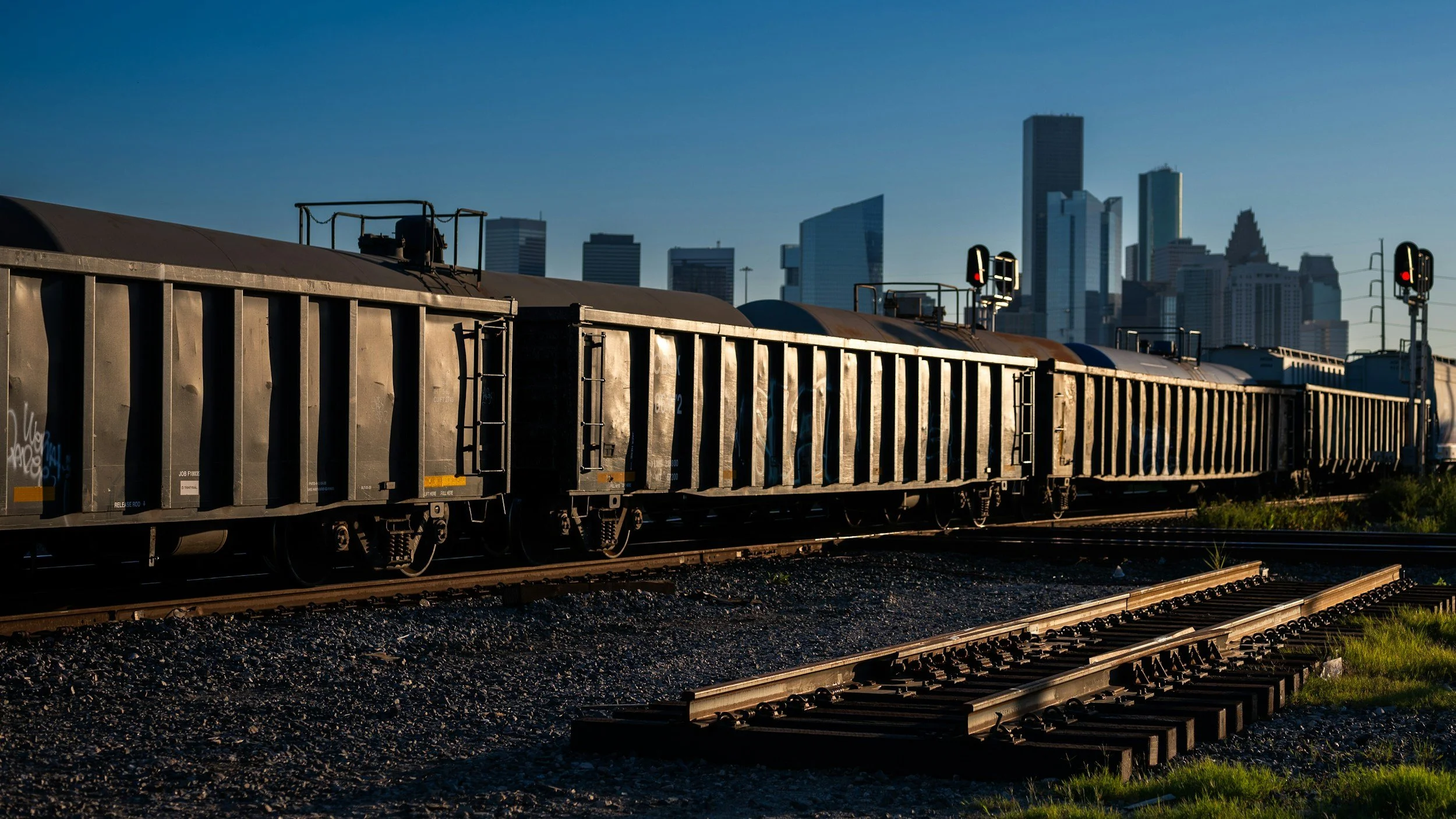 A freight train with black cars on railway tracks with a city skyline in the background, during daylight.