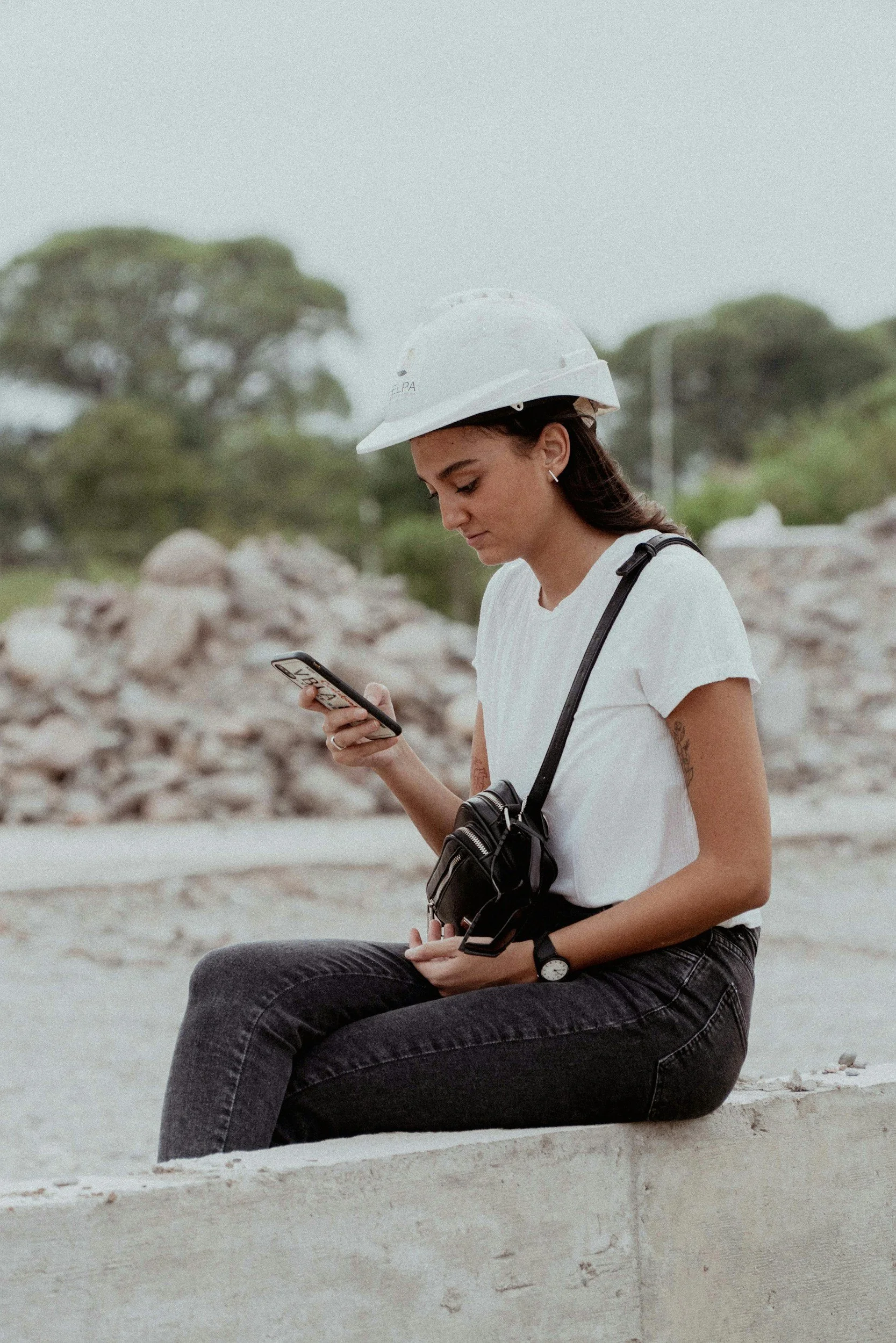 Jovem feminina sentada na beira de um muro, usando boné branco, camiseta branca, calça jeans escura, carregando uma câmera pendurada no pescoço, olhando para o celular, vestindo um relógio e brincos pequenos, ao fundo árvores e céu nublado.