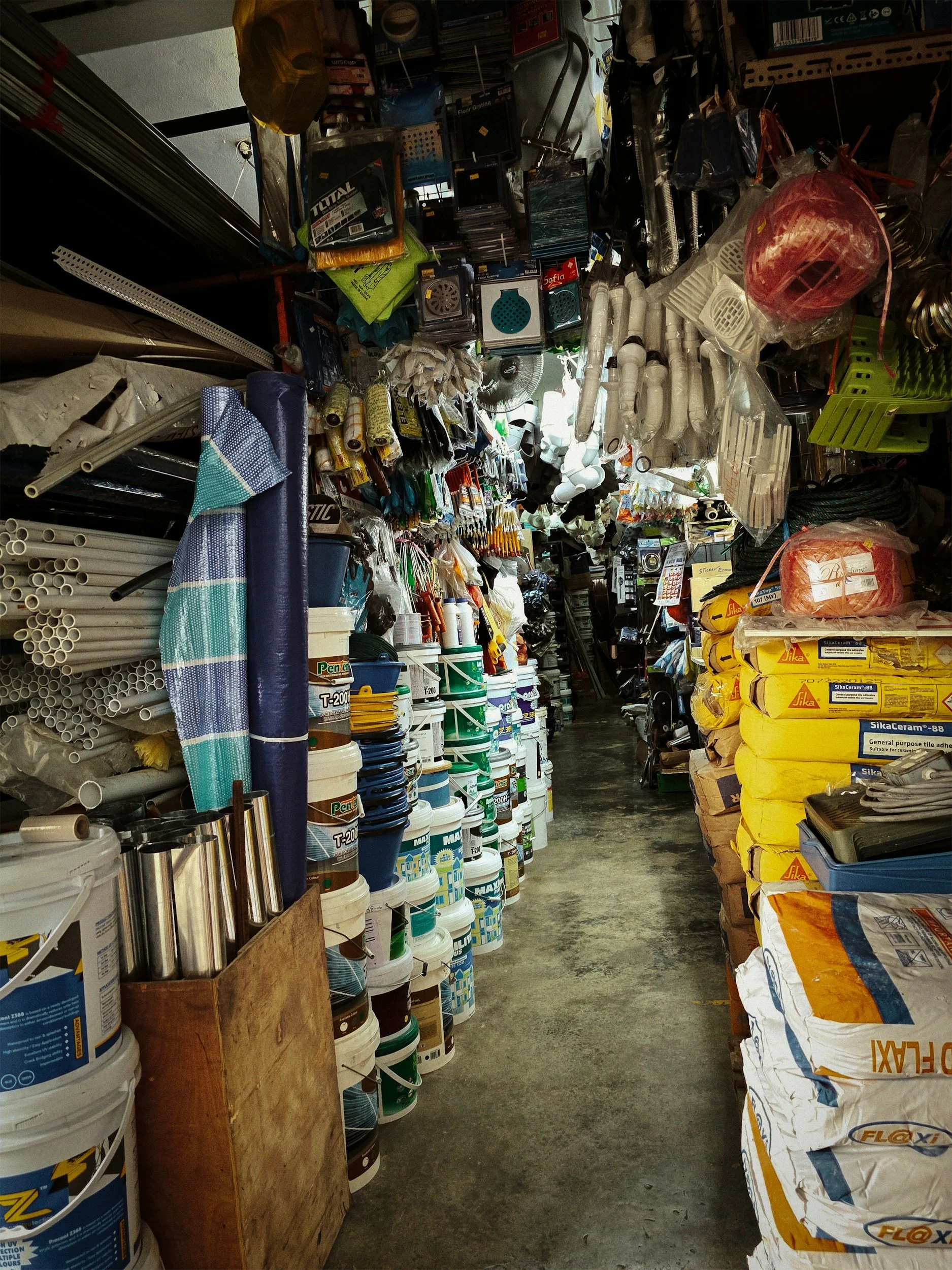 Imagem de uma loja de materiais de construção com prateleiras cheias de tintas, calhas, tubos de PVC, sacos de cimento e outros itens relacionados à construção civil.