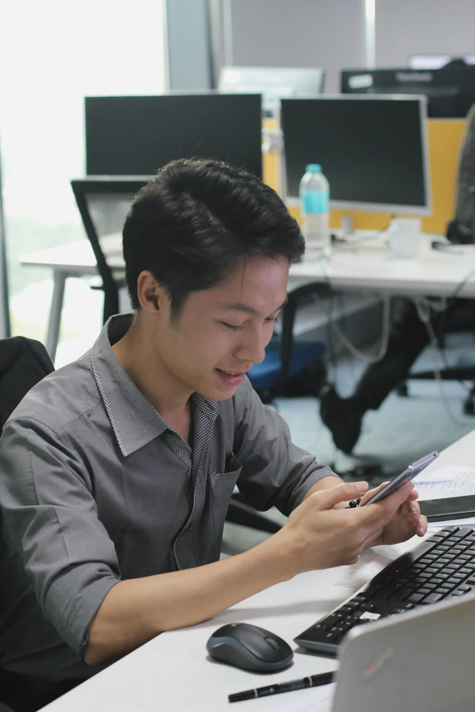 Homem sentado em uma mesa de escritório, usando camisa cinza, olhando para o celular, com teclado e mouse na frente e computadores ao fundo.