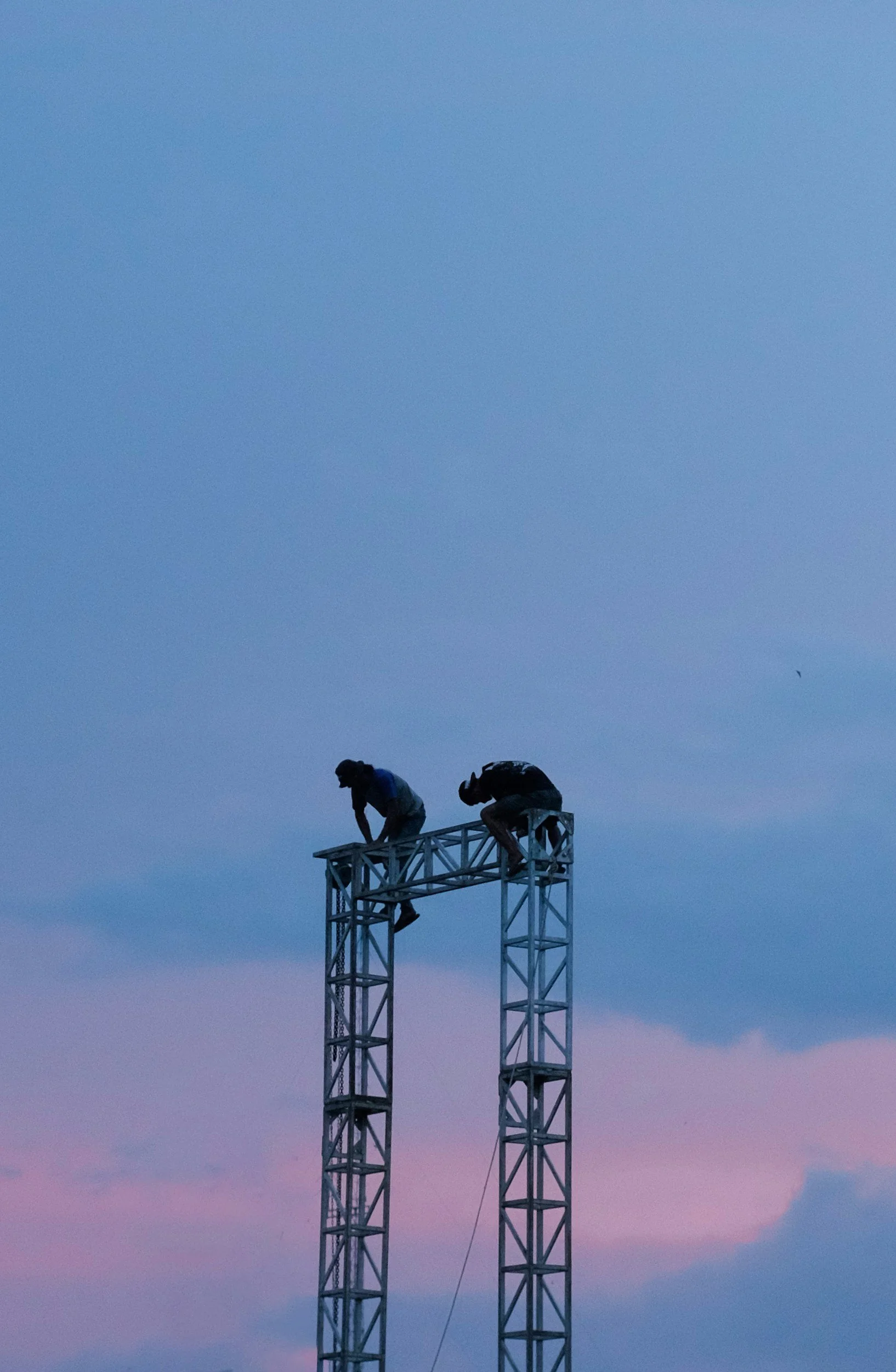 Dois homens trabalhando no topo de uma estrutura metálica durante o entardecer, sob um céu azul com tonalidades rosa.
