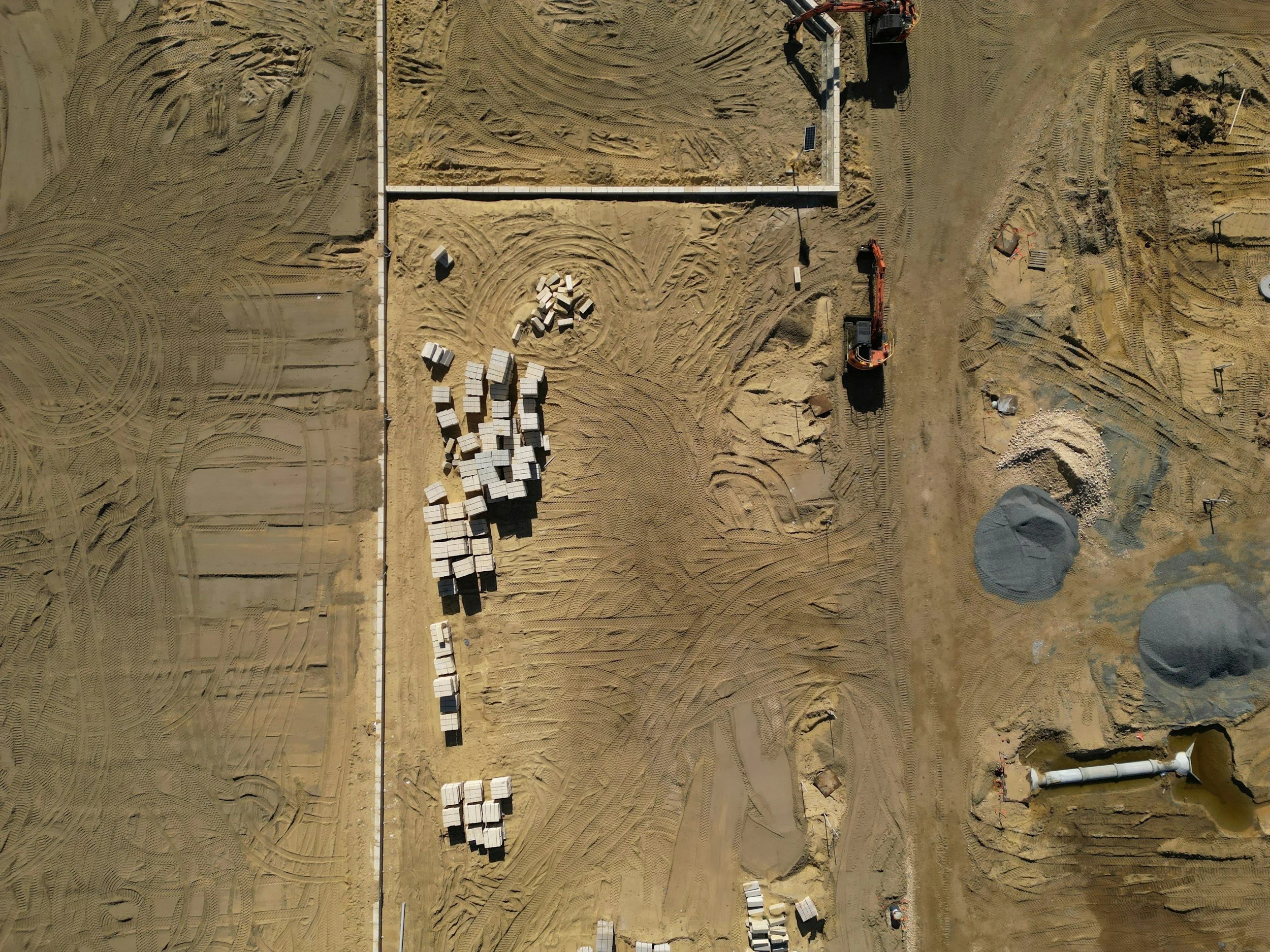 Imagem aérea de um canteiro de obra em construção com espalhamento de areia, blocos de construção empilhados, máquinas de escavação e instalações de energia solar.