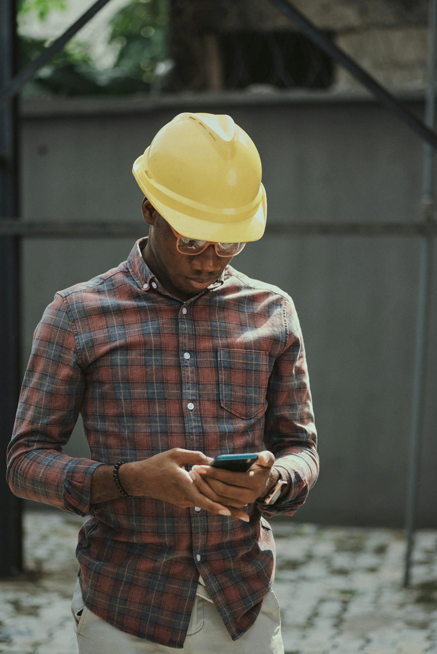 Homem com pele escura usando óculos de sol, camiseta de manga longa xadrez, com um boné de capacete amarelo na cabeça, segurando um celular e olhando para a tela, ao ar livre, com estrutura metálica e fundo de areia e vegetação.