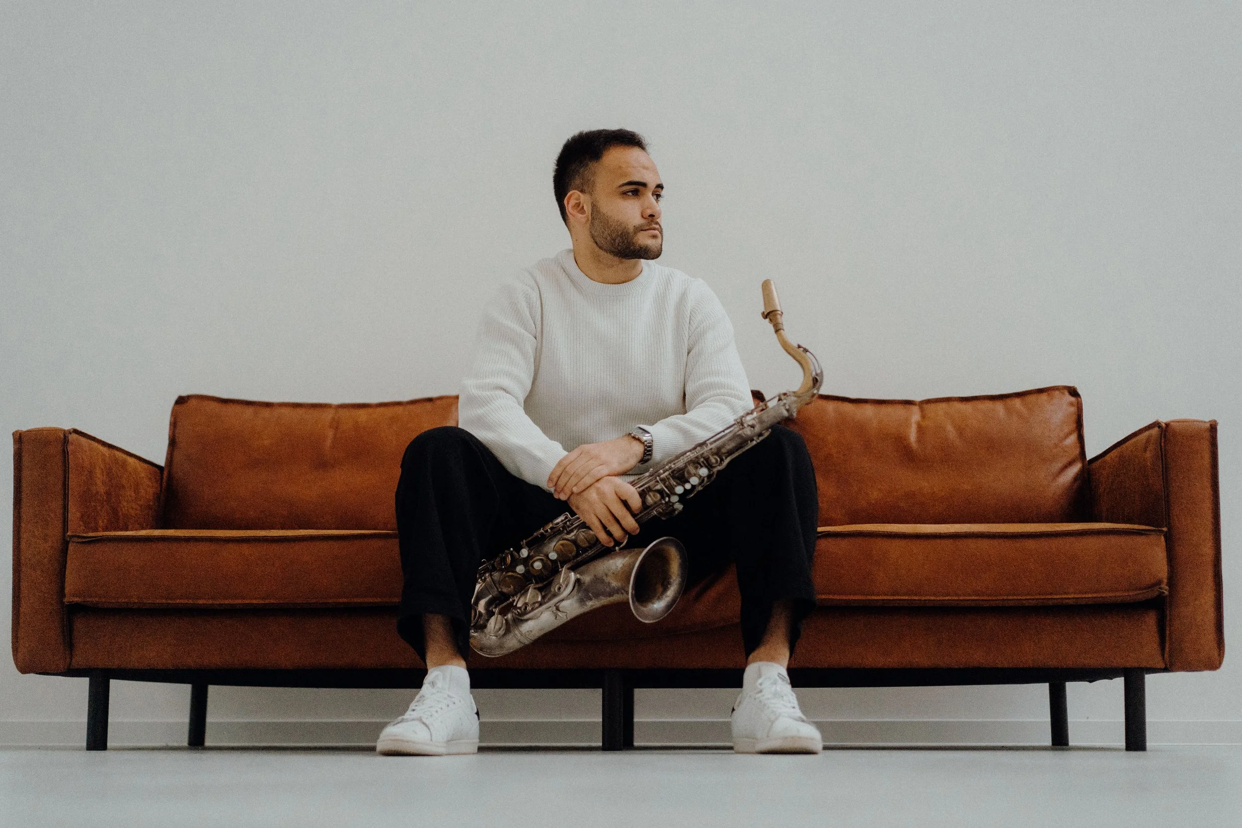 A teacher sitting on a brown leather sofa, holding a saxophone, wearing a white sweater, black pants, and white sneakers, in front of a plain white wall.