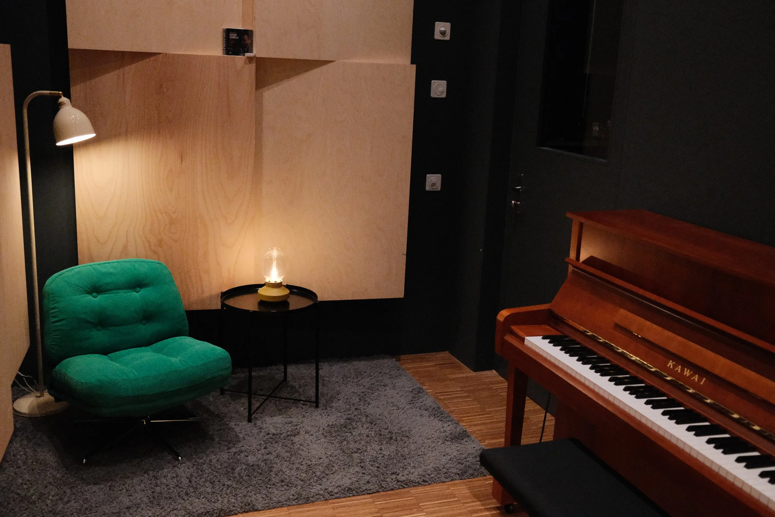 A cozy room with a green cushioned swivel chair, a small round black table with a lamp, a wooden piano, and a wooden wall with books. The lighting is warm and dim.