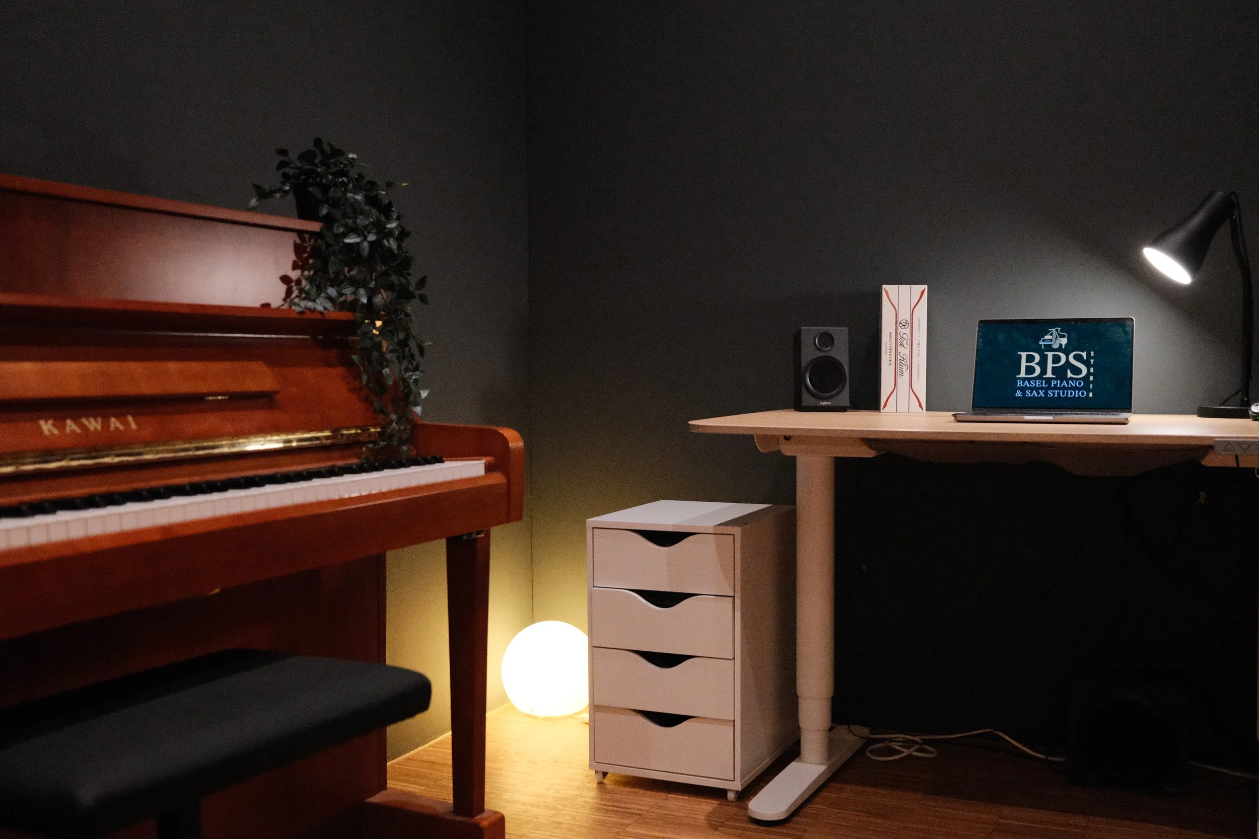 A cozy room featuring a wooden upright piano with a green plant on top, a small white drawer unit, a desk with a laptop displaying "BPS Basel Piano & Sax Studio," a speaker, a zebra-print box, a desk lamp, and a glowing spherical lamp on the floor, all set against dark-colored walls.
