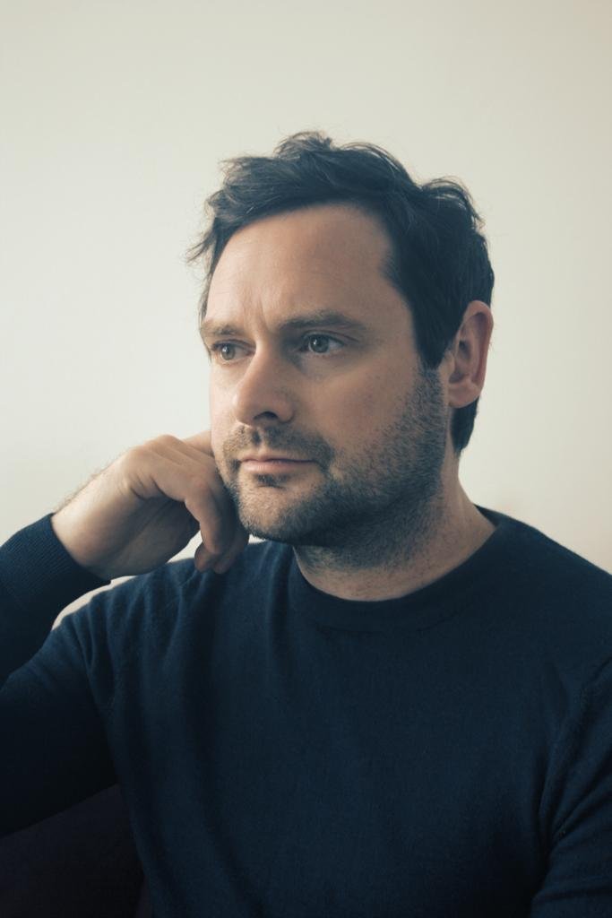 A man with dark hair, wearing a black shirt, sitting against a light-colored wall, resting his chin on his hand and looking thoughtfully to the side.