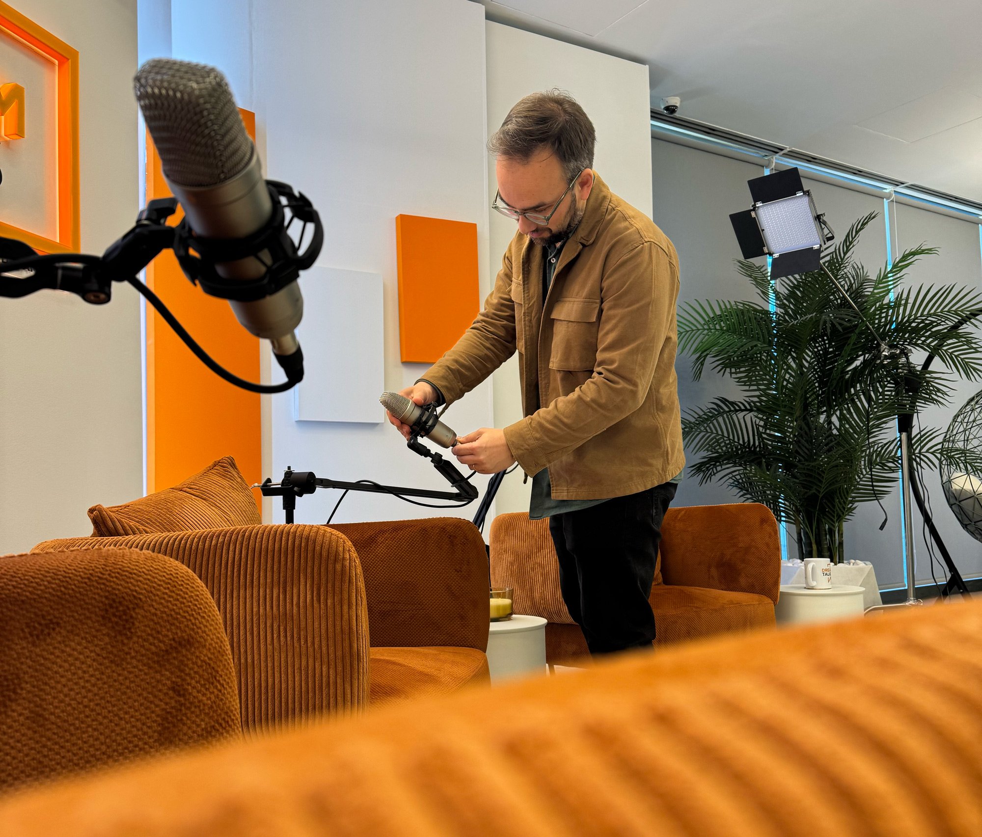 Homem ajustando equipamento de gravação em ambiente de estúdio ou sala de reunião com sofá laranja, plantas e iluminação profissional.