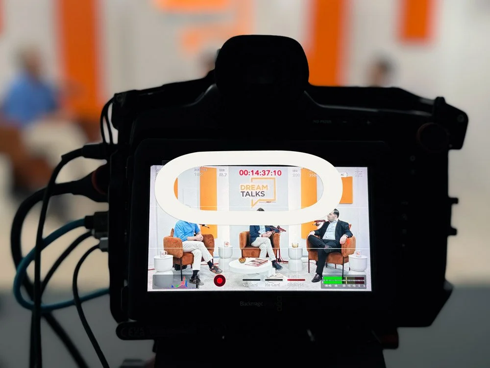 Câmera filmando uma discussão ao vivo de três pessoas em um set de gravação de programa de TV, com o fundo do estúdio visível