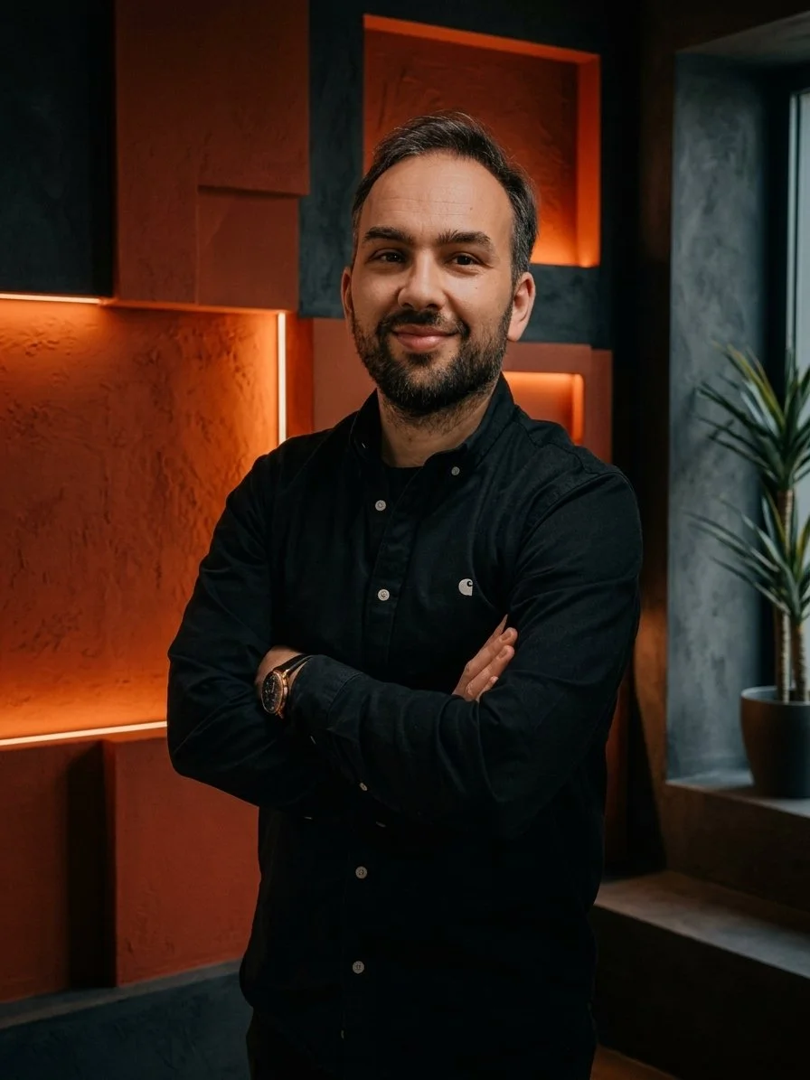 Homem sorridente com barba, cabelo castanho, usando camisa preta, posando com braços cruzados em um ambiente com iluminação amarelada e paredes coloridas, ao lado de uma planta.