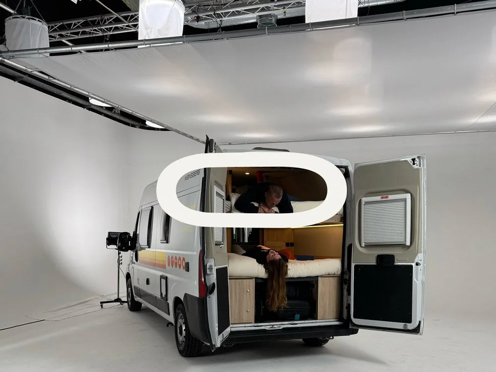 Interior de uma van adaptada com cama, com duas pessoas conversando, uma de cabeça para baixo na cama e outra de pé, em um ambiente de estúdio fotográfico.
