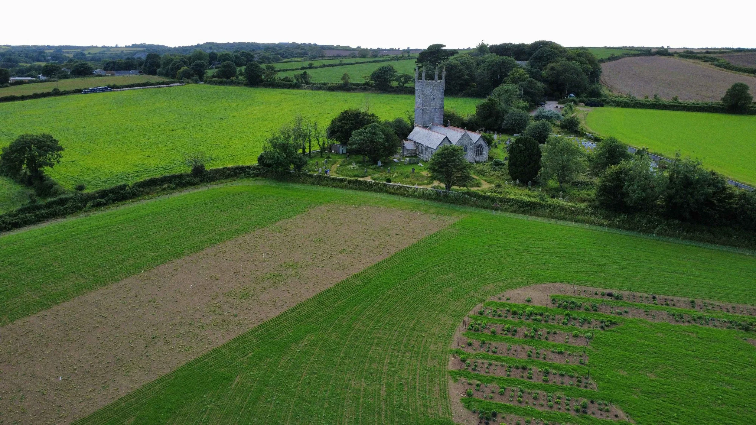 Aerial view of a rural landscape with green fields, trees, and an old church with a tall stone tower.