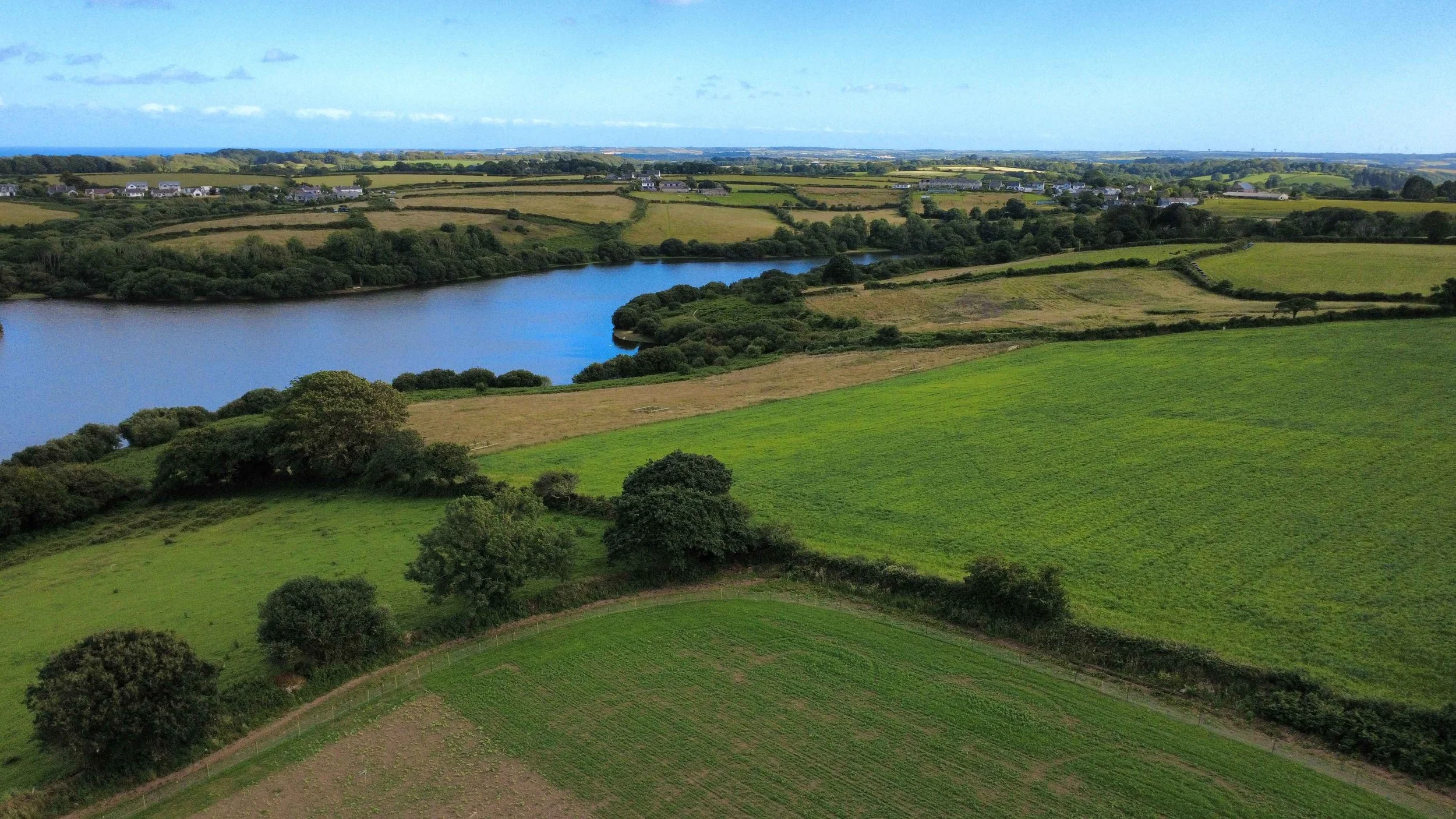 A scenic landscape with rolling green fields, a reservoir, and small clusters of trees and bushes, with houses and fields in the background under a partly cloudy sky.