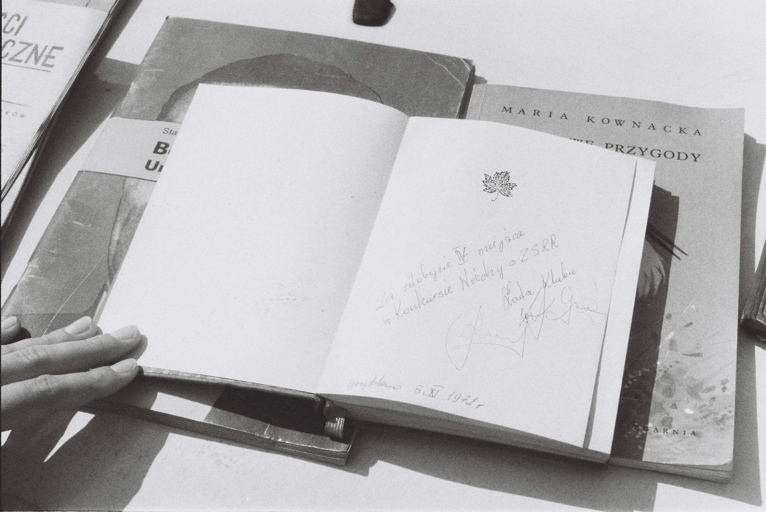 A person's hand holding an open book or notebook on a table, with handwritten notes visible. Several other books or materials are also on the table, including one titled 'Maria Kownacka' and another with a partially visible word 'Grazka.' The photo is in black and white.