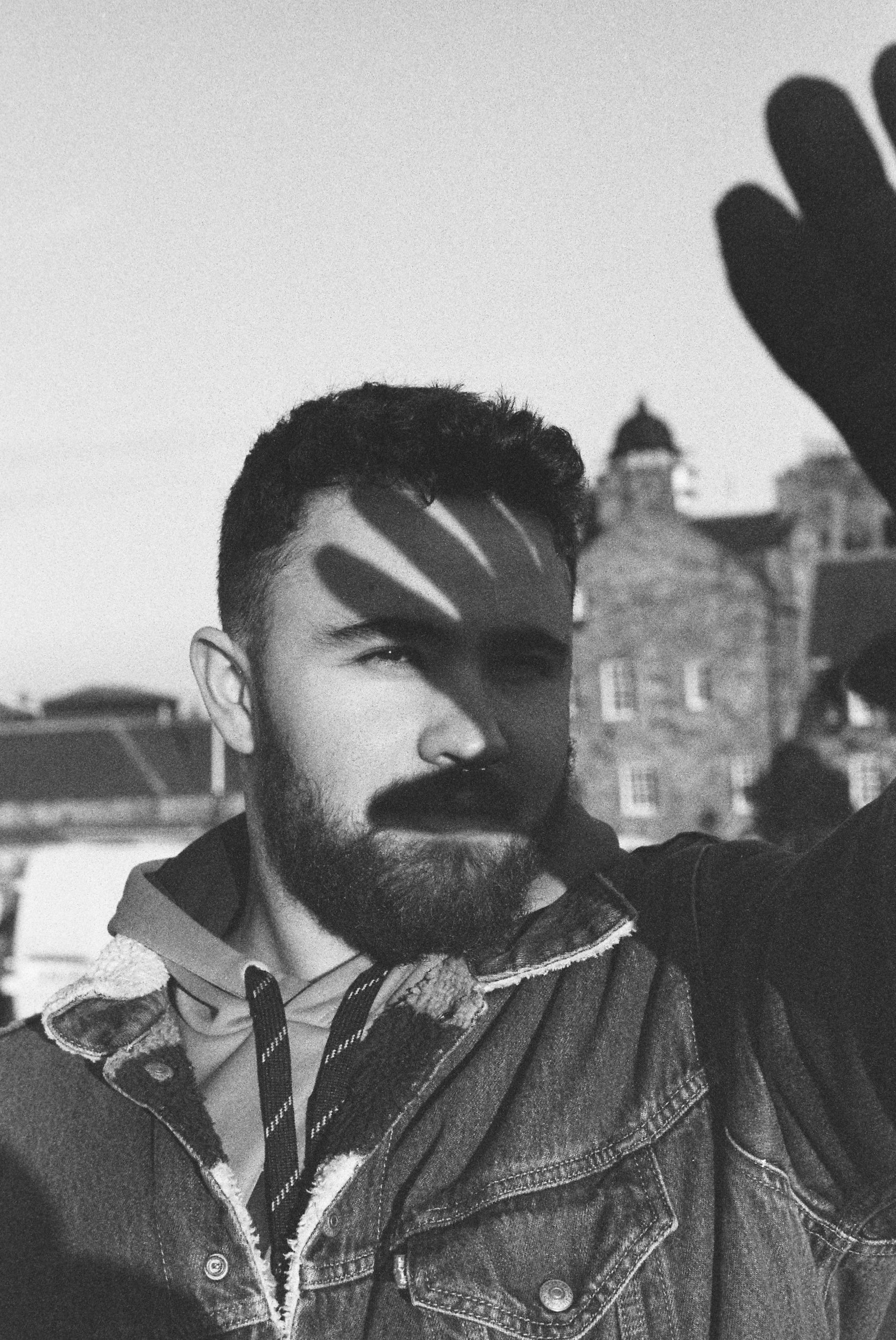 A black and white photo of a young man with a beard and short hair, wearing a denim jacket and a hoodie, with buildings and a dome in the background, raising his hand.