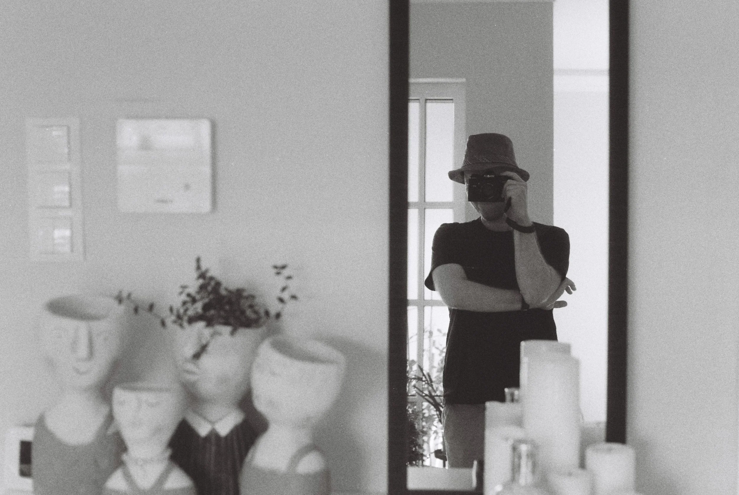 A person wearing a bucket hat taking a photo of their reflection in a mirror, with a window in the background and a plant in the foreground.