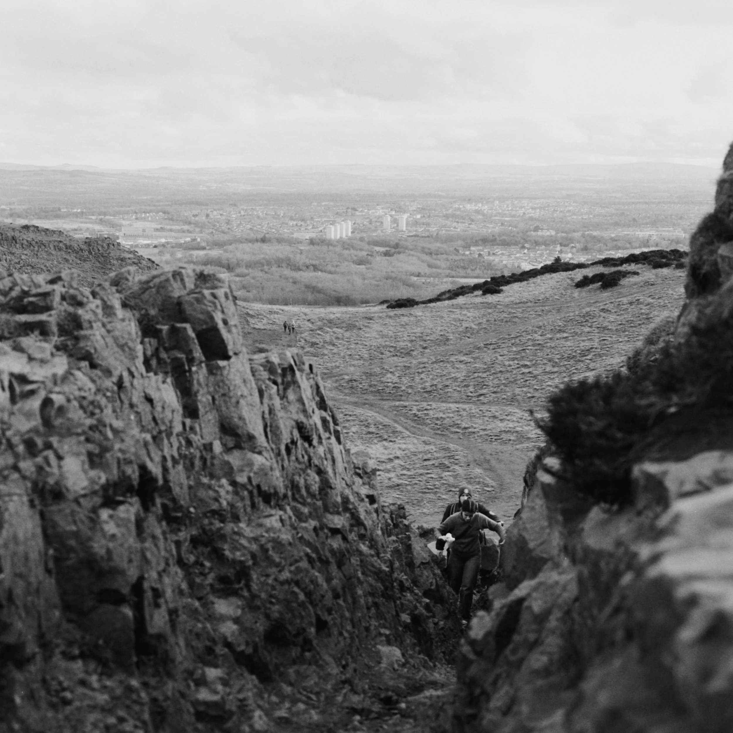 A black-and-white photograph of a wide landscape view taken from an elevated position. The foreground shows a rocky terrain with a few people walking in the dirt path. The middle ground has a few trees and low hills, and the background features a cityscape with tall buildings and a distant horizon under a cloudy sky.