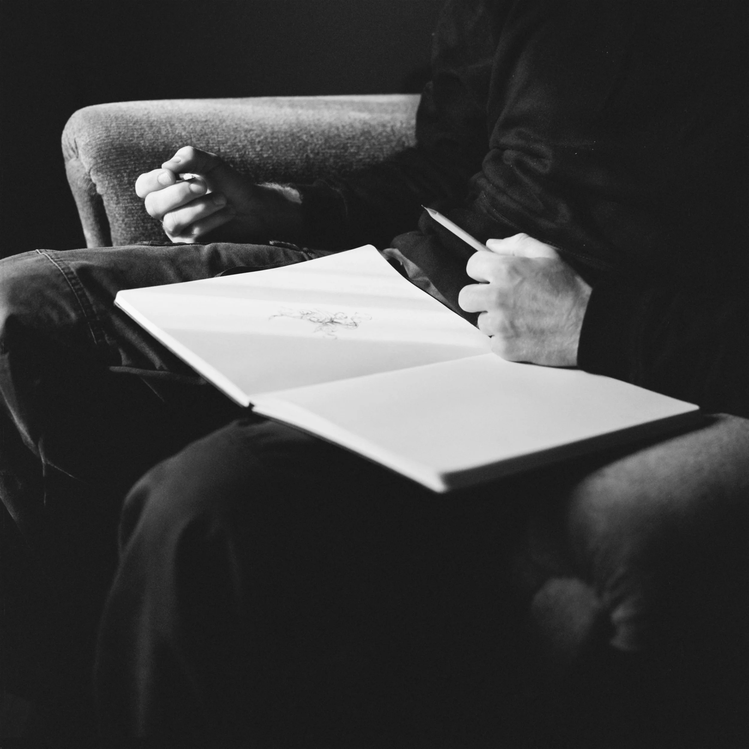 A person seated on a sofa holding a smartphone in their left hand and a notebook or paper in their right hand, with a pencil resting on the notebook. The person is wearing a dark jacket and jeans. The focus is on the hands and the objects they are holding.