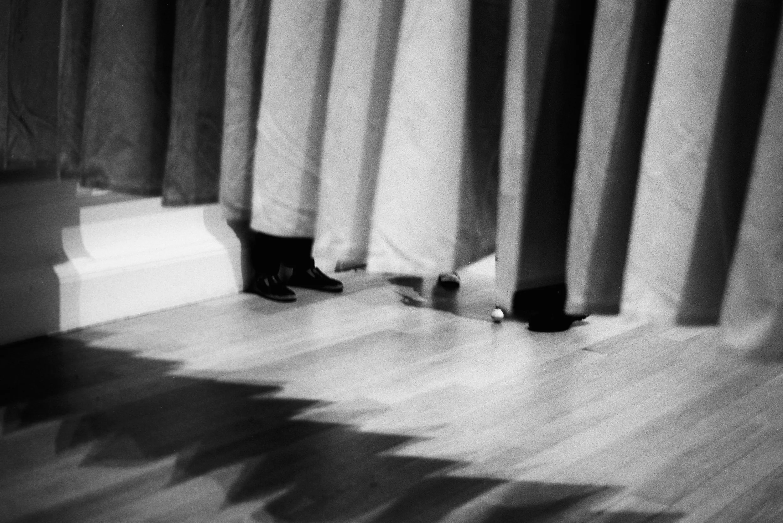 Black and white photo showing the lower legs and feet of a person standing on a wooden floor, with another person's feet visible in the background. The scene appears to be viewed through a series of vertical blinds or bars.
