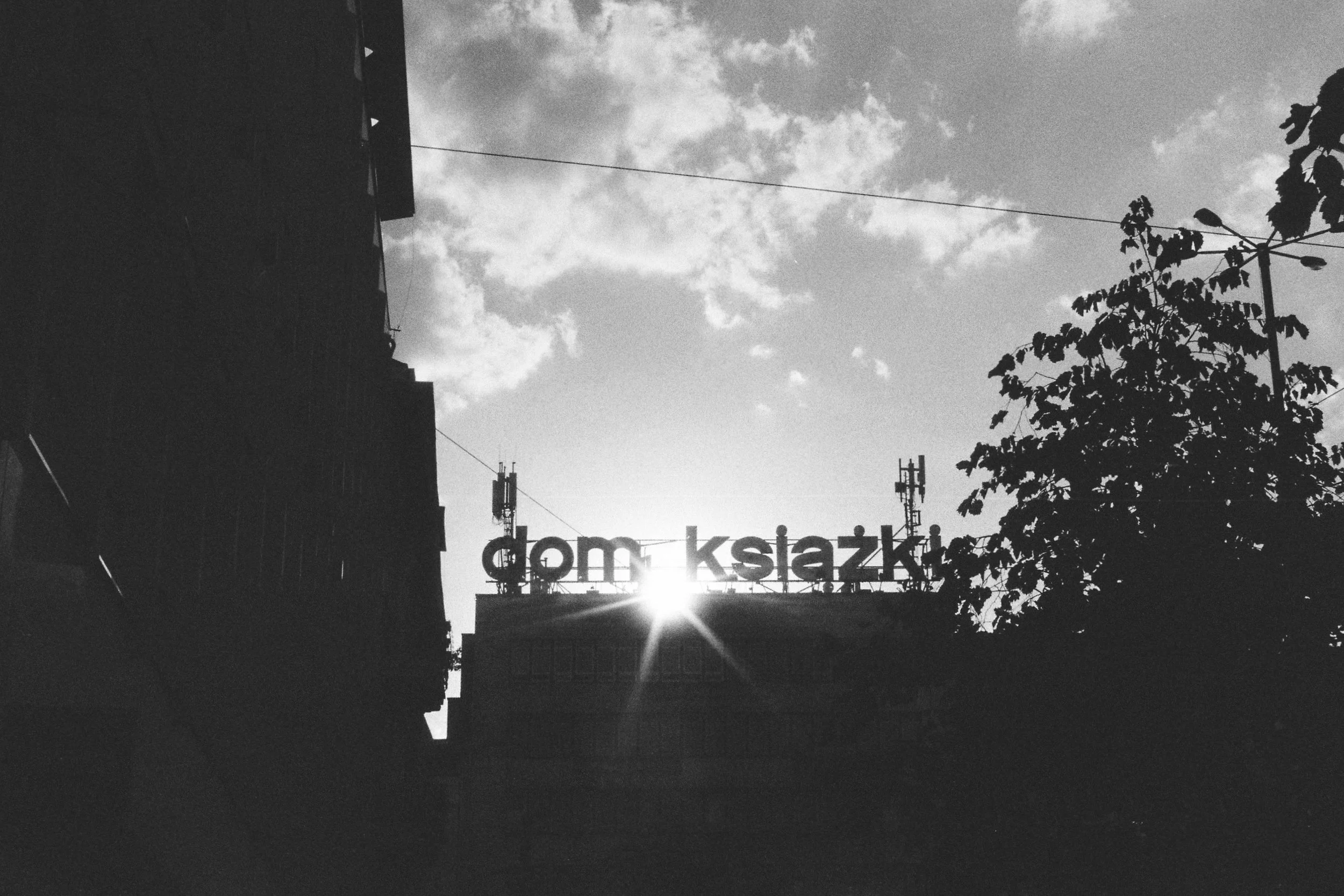 Black and white photo of city buildings with a sign that reads 'dom ksia' in the background, a tree on the right, and clouds in the sky.