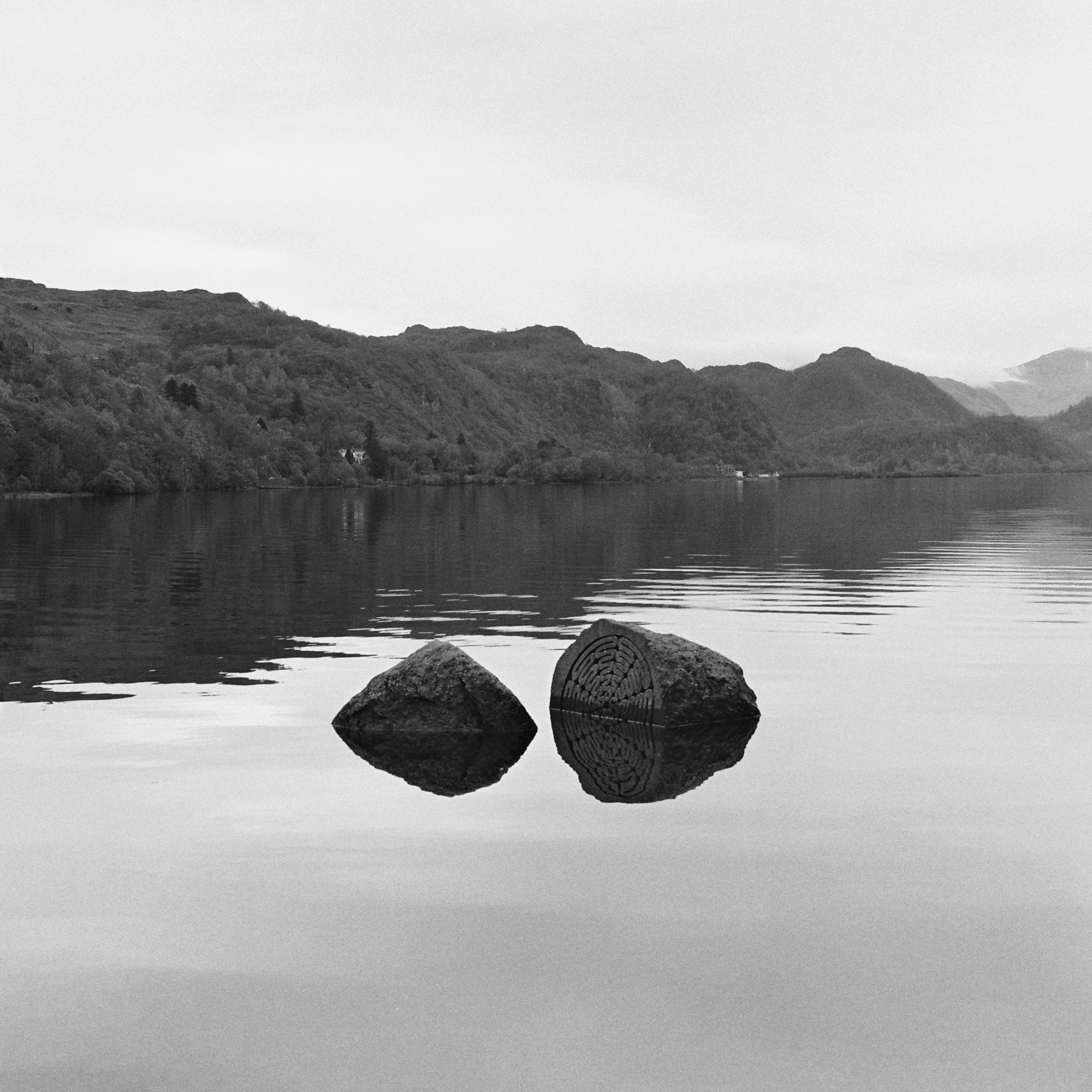 Black and white photo of carved rocks in the foreground on a calm body of water with a mountain range in the background under a cloudy sky.