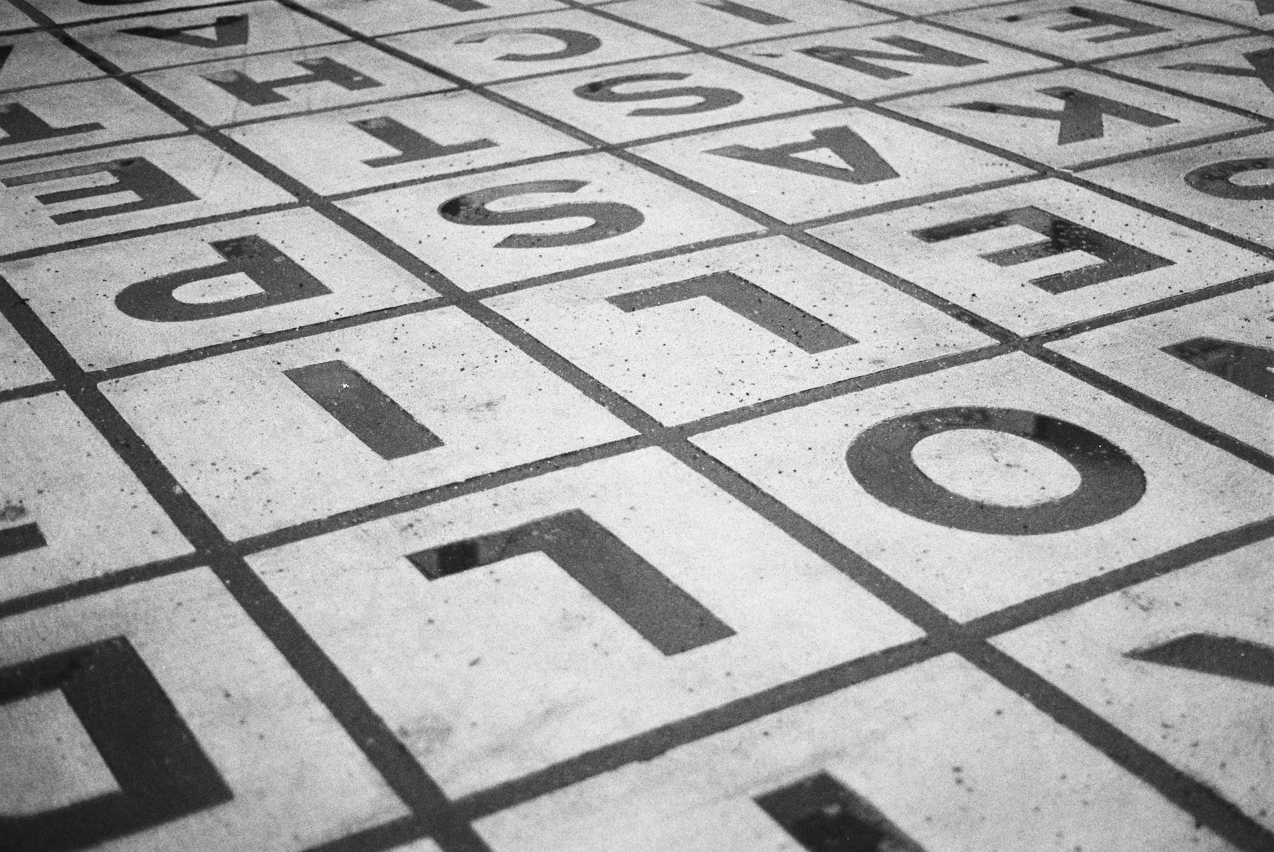 Black and white photo of a tiled floor with alphabet letters and numbers in a grid, resembling a hopscotch pattern.