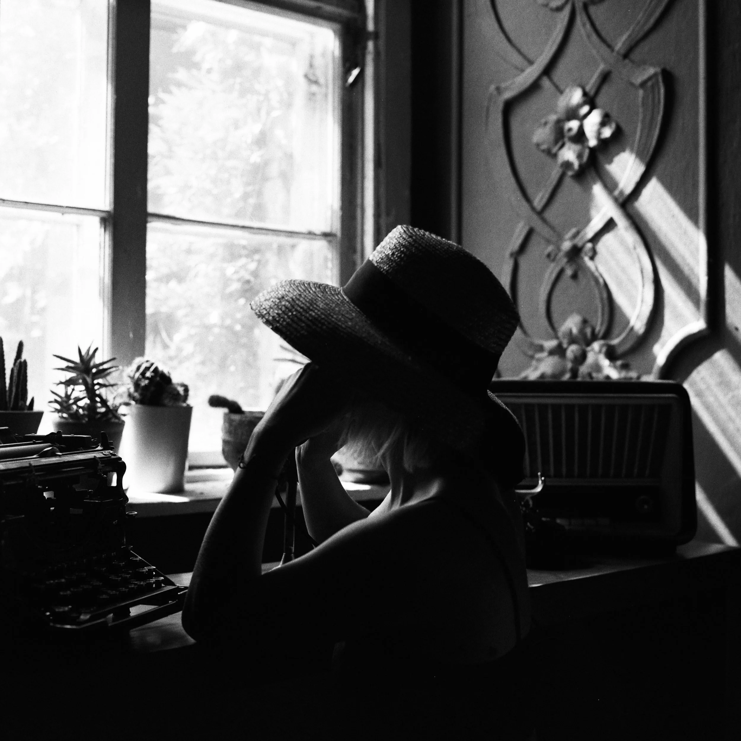 Black and white photograph of a window with light coming through, plants on a ledge, a radiator, and a person with legs crossed, wearing a knitted sock or glove near the window.