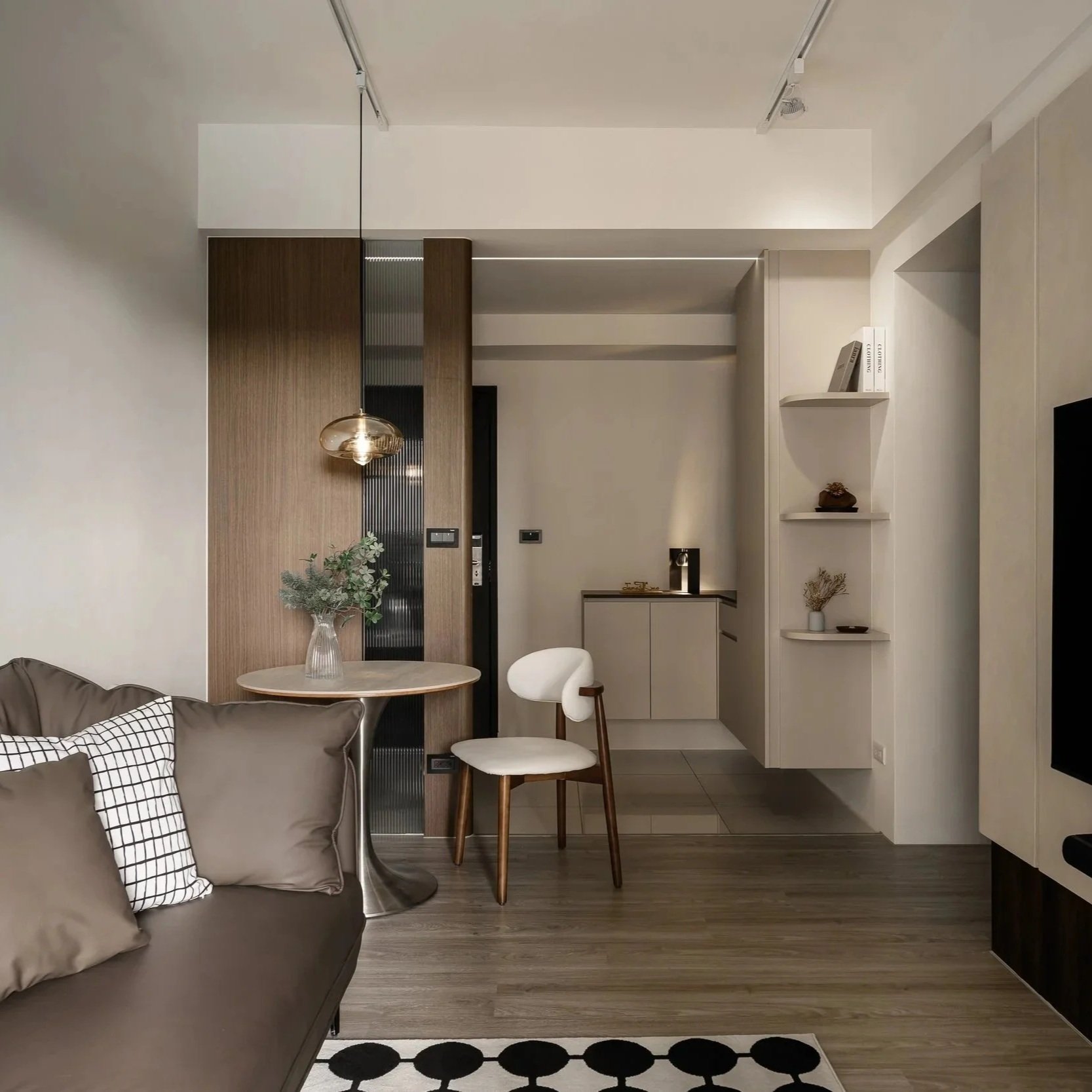 Interior of a modern apartment with a round dining table, beige sofa with pillows, a decorative vase of greenery, a white chair, a small kitchenette area, and a built-in shelf with books and decor.