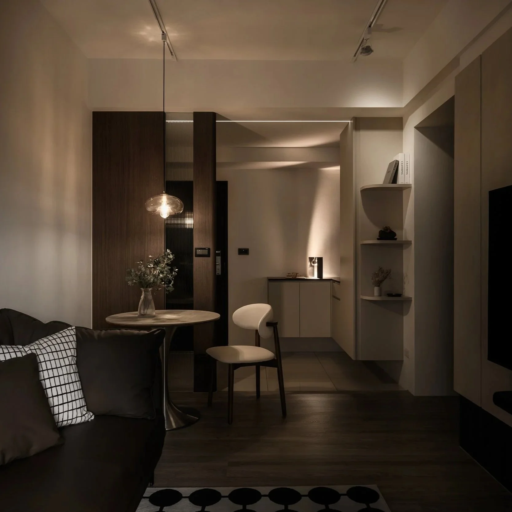 Living room with dark sofa and checkered cushions, small round dining table with a glass vase of flowers, two chairs, kitchen area with cabinets, open shelving with books and decorations, warm lighting creating a cozy atmosphere.