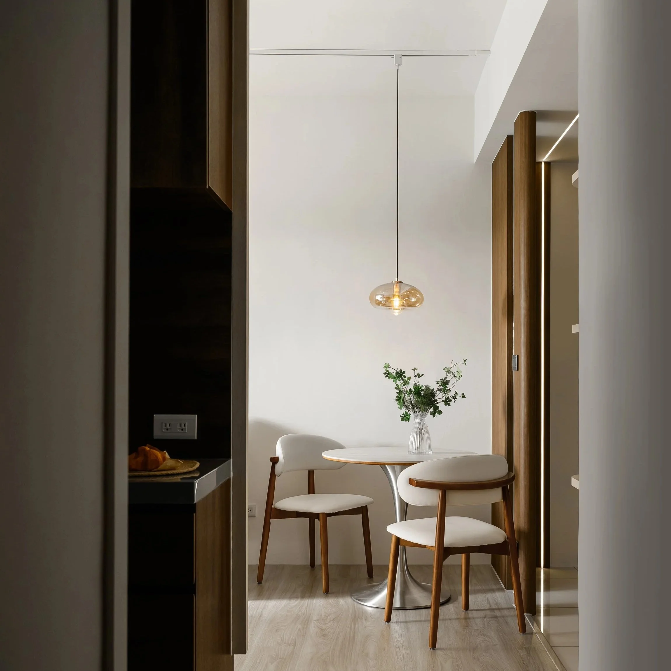 A dining area with a round table, two white chairs with wooden legs, a clear vase with green plants, and a hanging pendant light in a modern, minimalistic space.