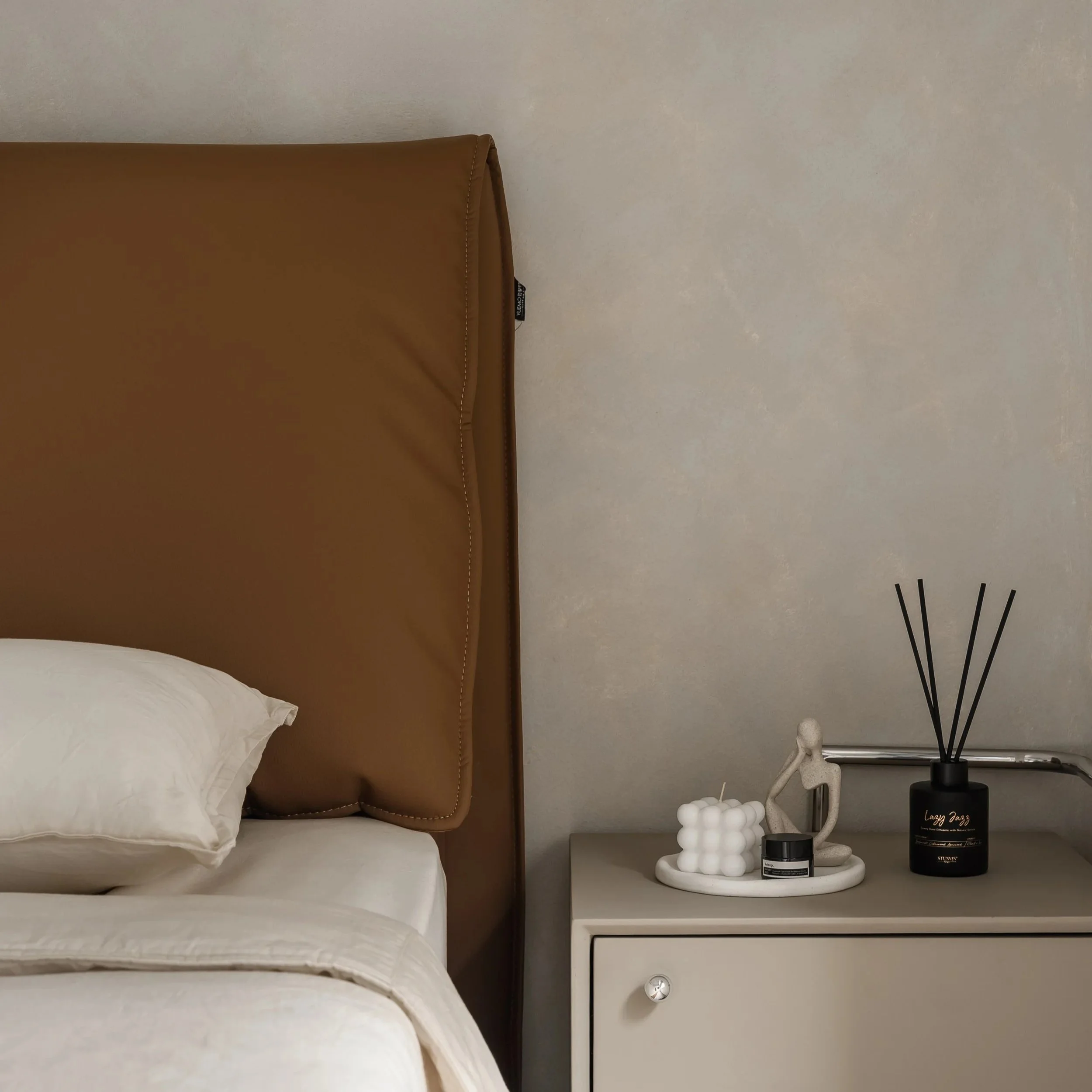 A section of a bedroom with a white bed and a brown leather headboard, a white nightstand, and a modern black lamp with white spherical accents. The wall behind is plain and light-colored, and there is a small black sign on the nightstand.