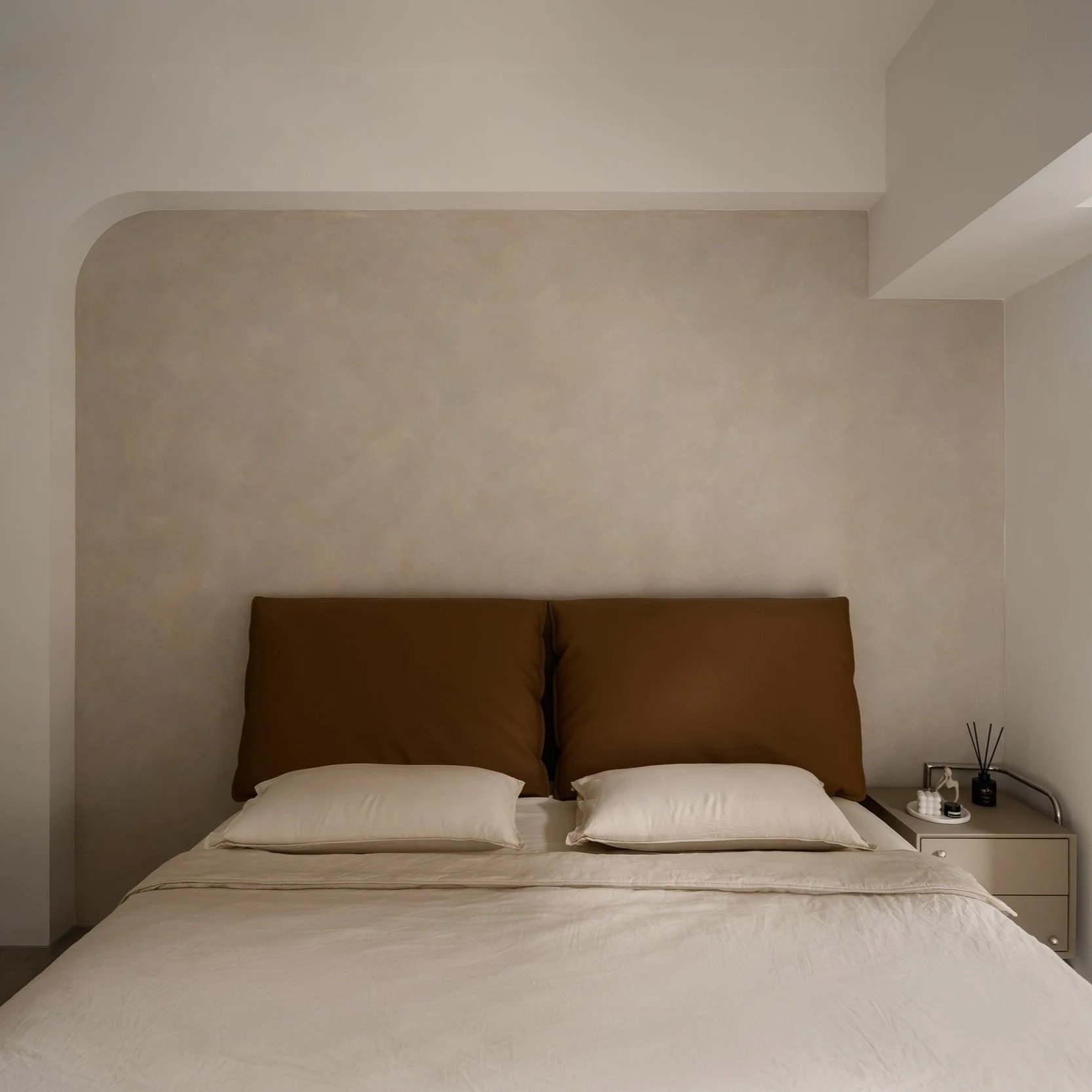 A neatly made bed with white sheets, beige pillows, and a brown headboard in a minimalist bedroom. A small nightstand holds a black reed diffuser and decorative items. The wall above the bed is plain with subtle texture.