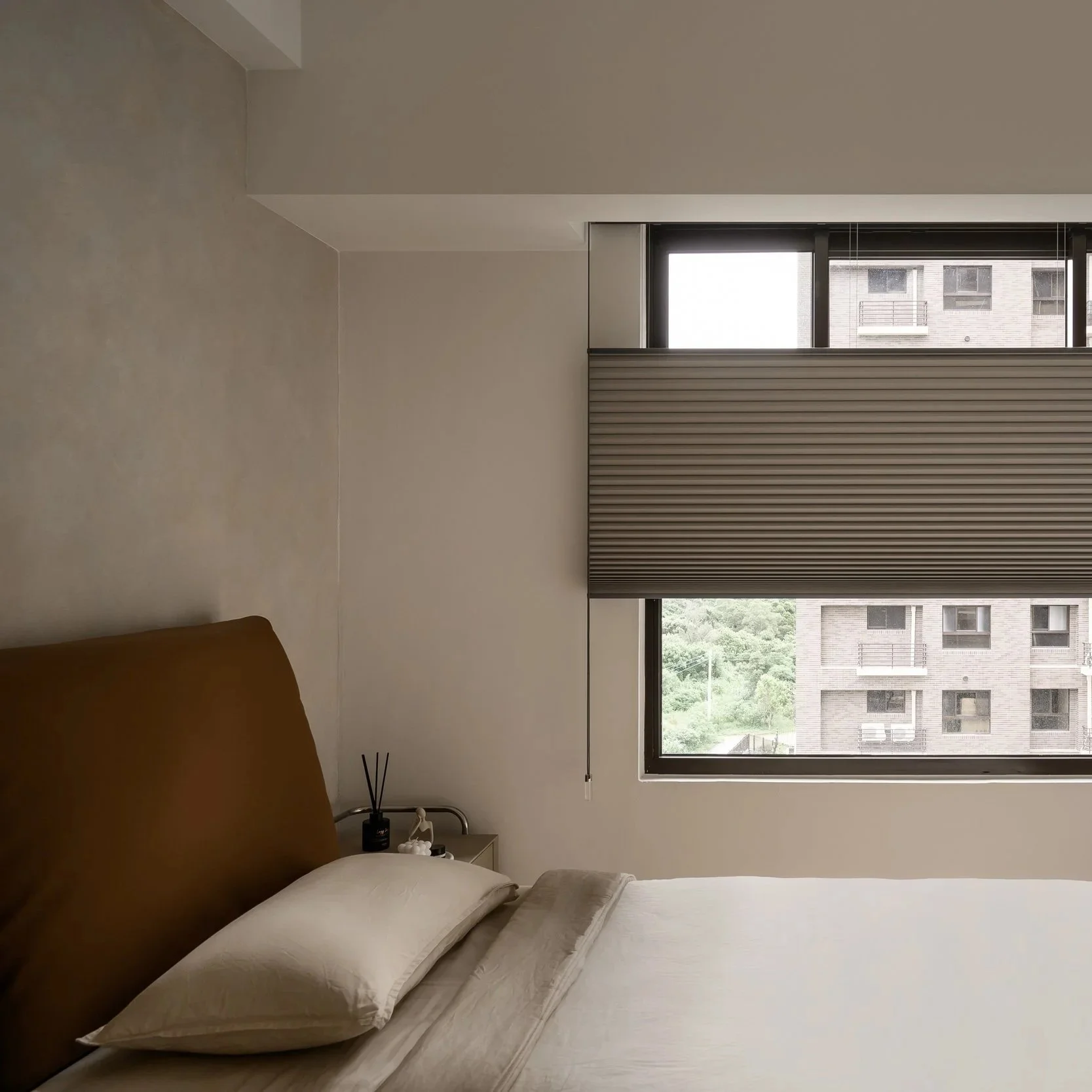 A bedroom with a bed, pillows, and a window with blinds showing a building outside.