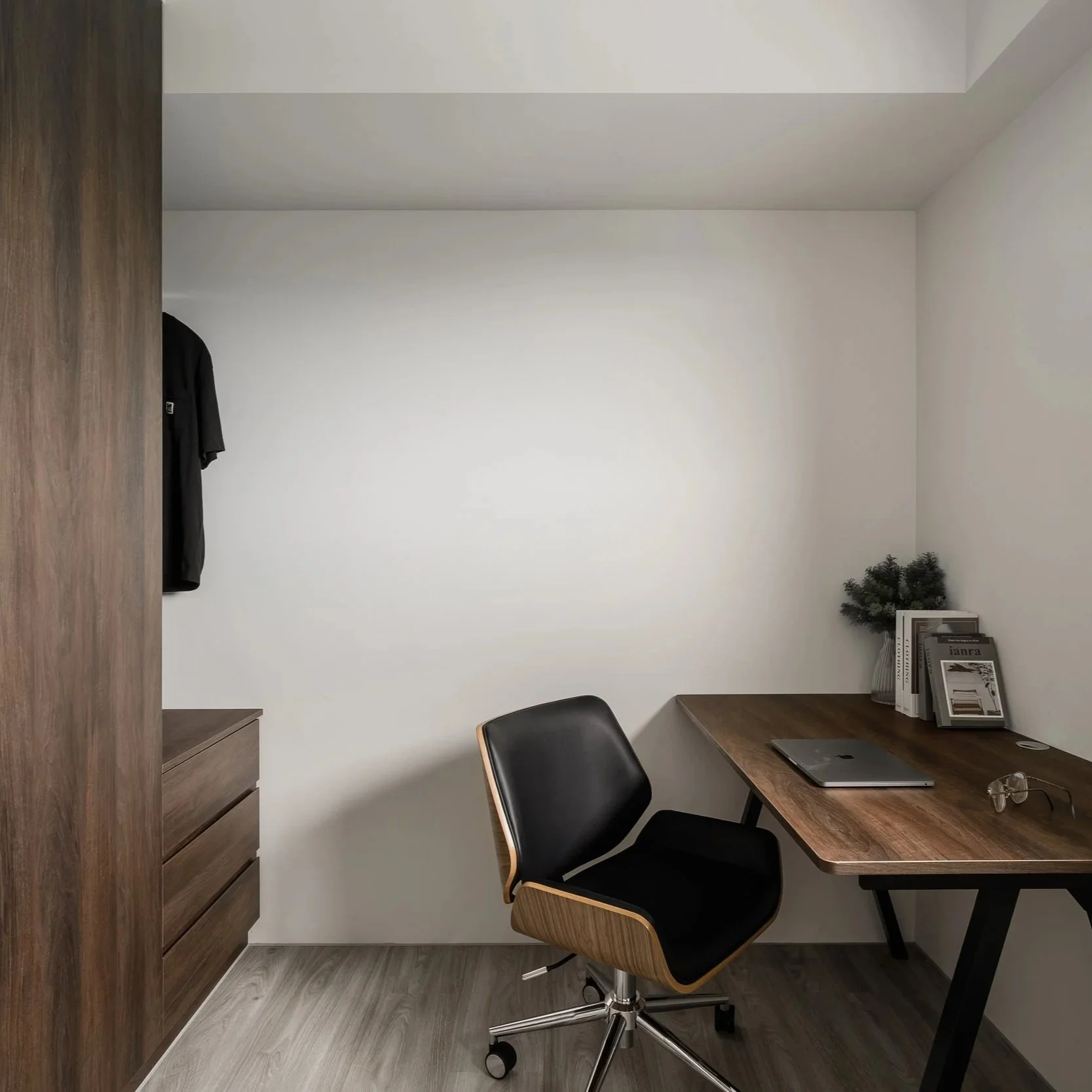 Modern minimalist office corner with wooden desk, black office chair, bookshelf, magazines, and minimal decor.