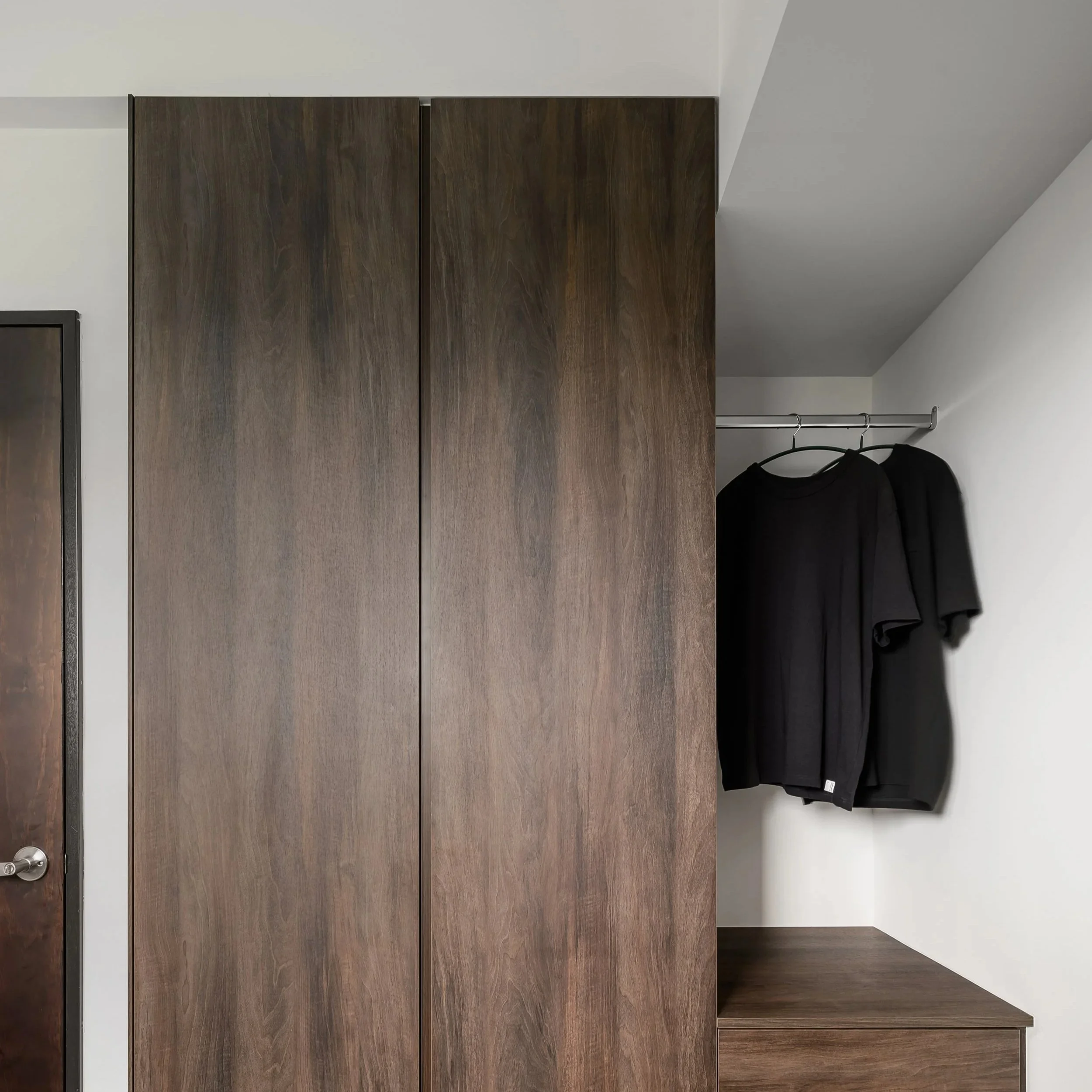 A wooden wardrobe with two doors and a small open section at the bottom, next to a closet rod with hanging clothes, against white walls.