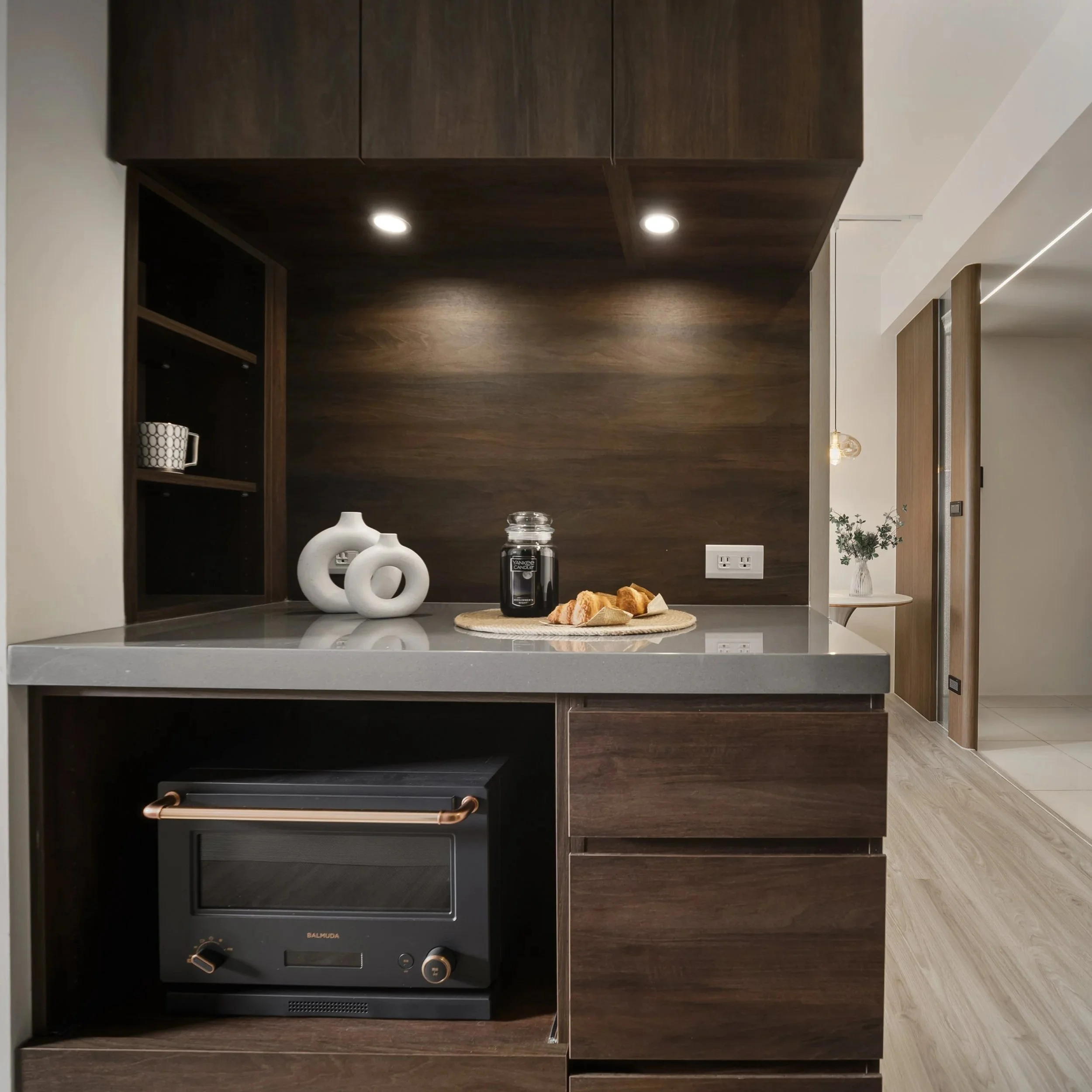 Modern kitchen with dark wood cabinets, two white decorative vases, a jar of dark chips, and slices of bread on a round woven placemat on a gray countertop under built-in ceiling lights.