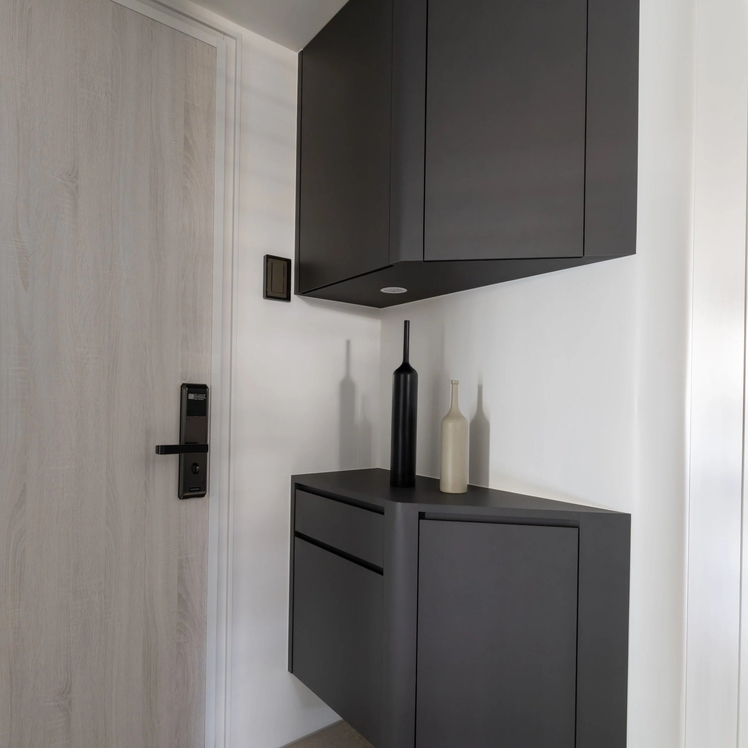 Interior view of a modern living space with a light wood floor, white walls, and black cabinetry. There are two white vases on a black shelf, a black table lamp, and an electrical outlet.