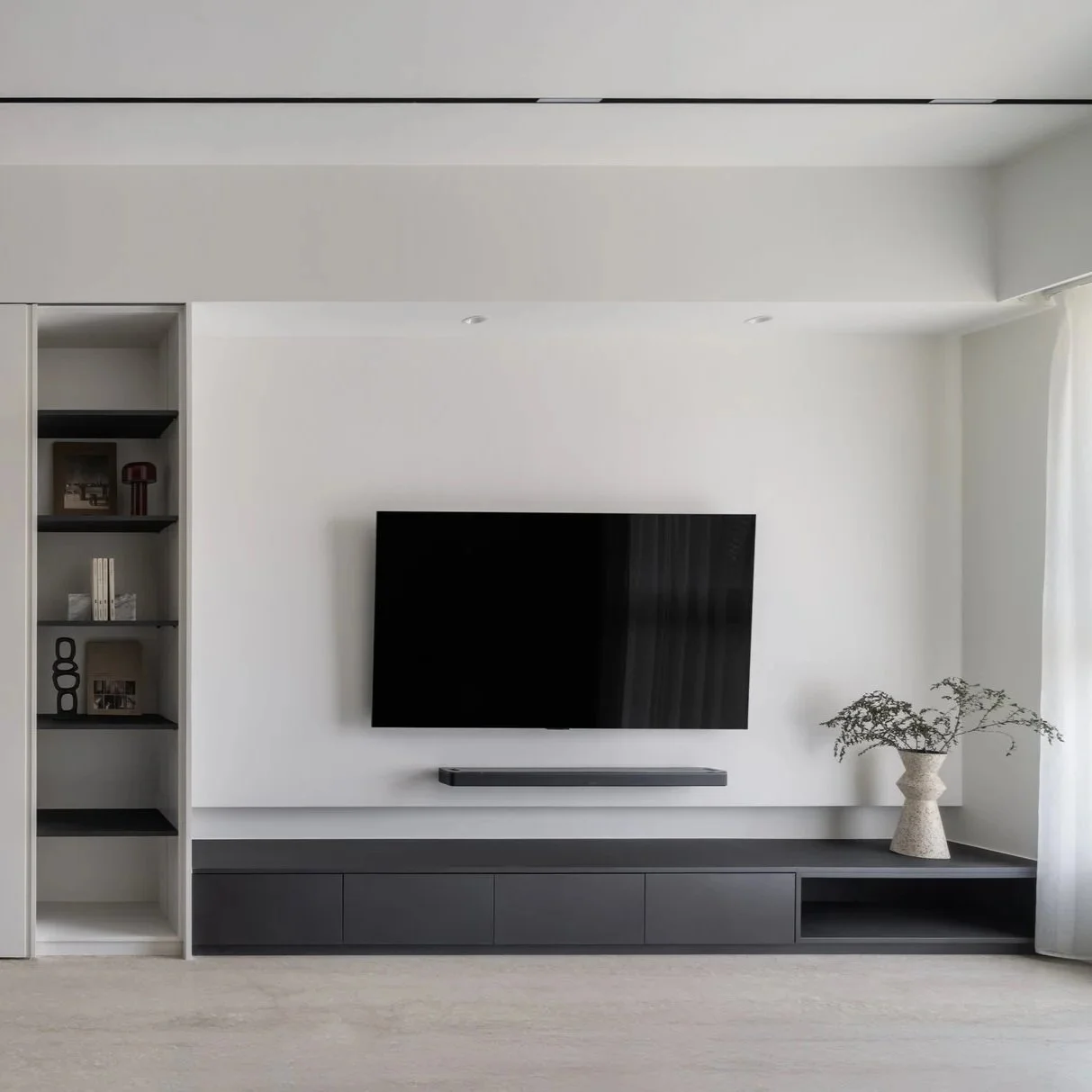 Minimalist living room with a wall-mounted flat-screen TV, a black soundbar below it, a built-in black TV stand with closed storage, and a large vase with greenery on the right side, all against white walls and ceiling.
