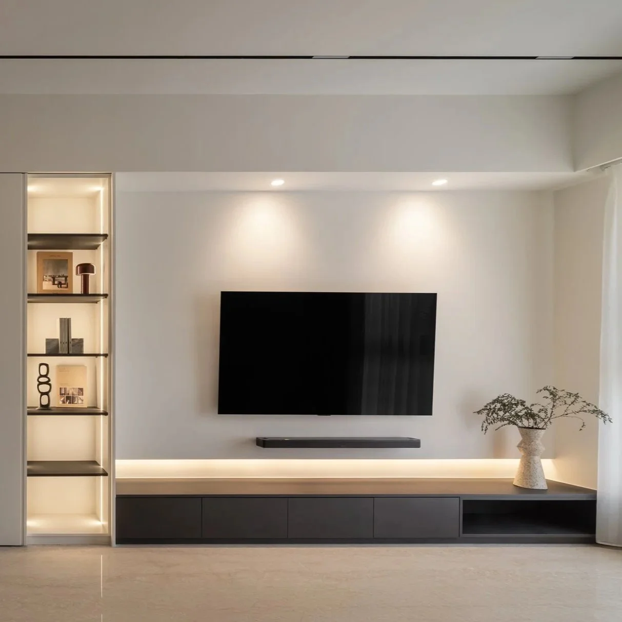 Modern living room with a wall-mounted TV, a black soundbar beneath it, built-in shelving with decorative items, and a vase with plants on a low cabinet, illuminated by recessed lighting.