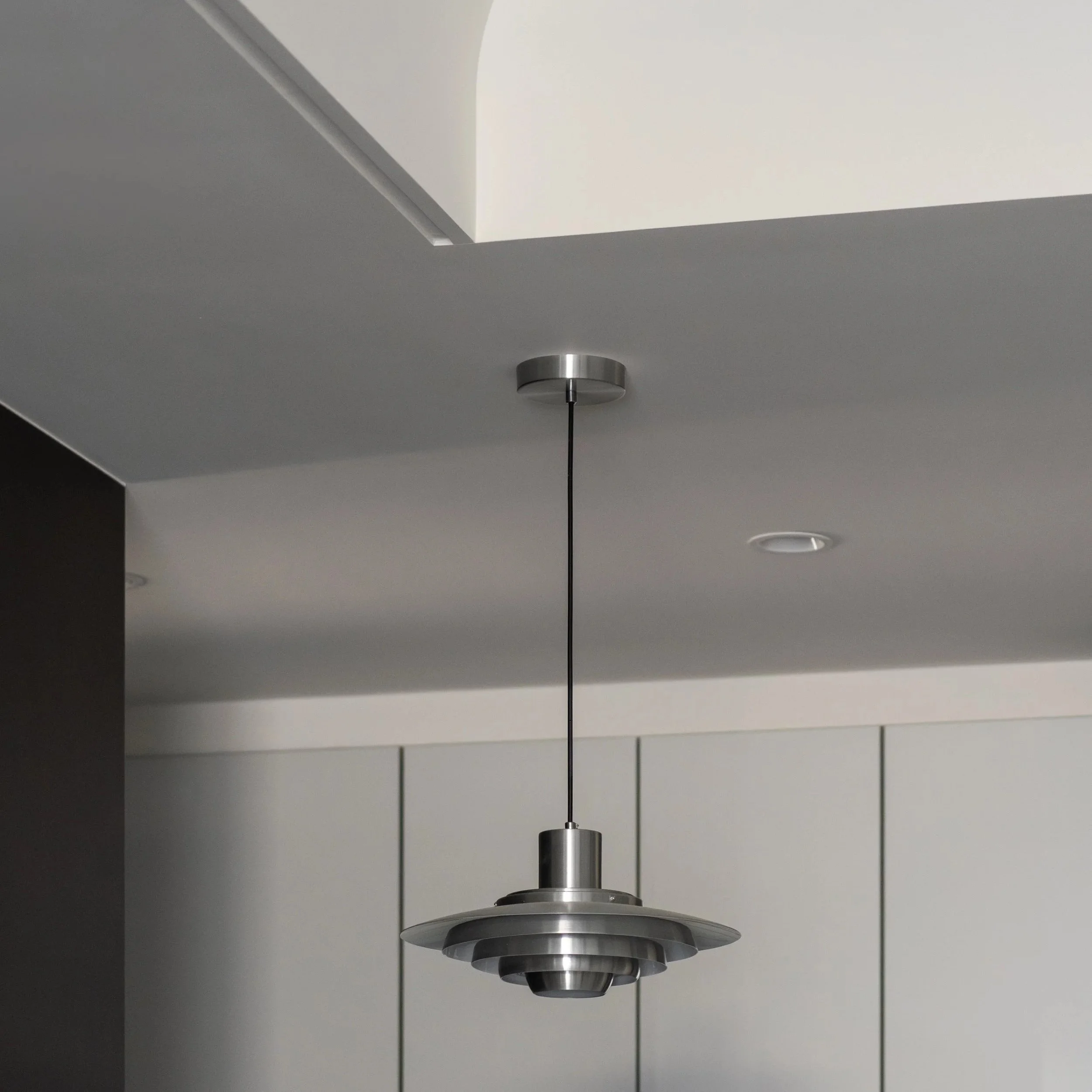 A modern hanging ceiling light fixture with multiple concentric metallic rings hanging from a black cord in a room with a white ceiling and gray walls.