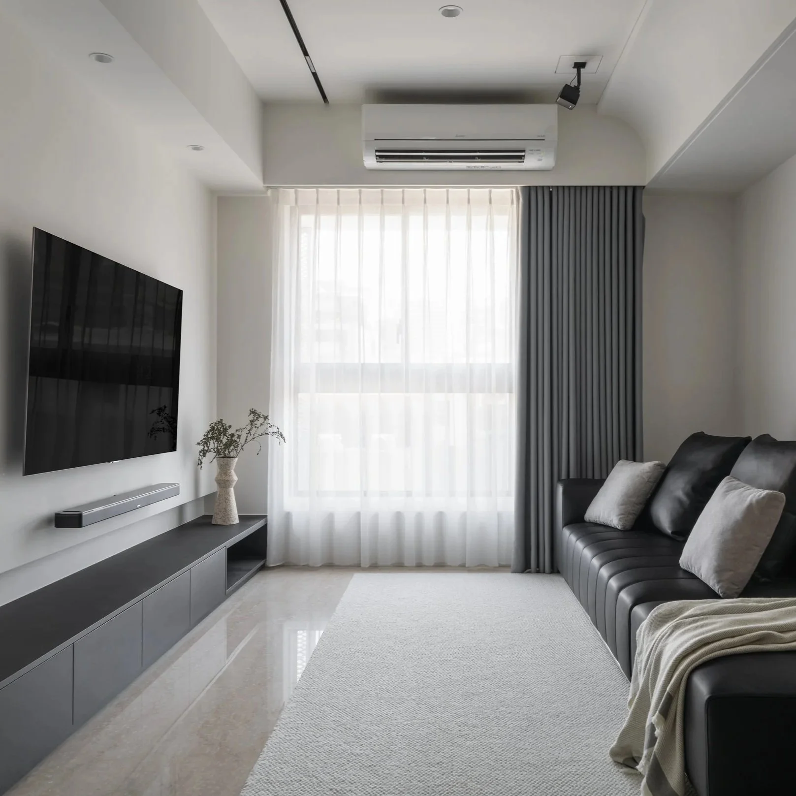 Modern living room with a black leather sofa with gray pillows, a large flat-screen TV on the wall, a white media console, a potted plant, and curtains covering a bright window.