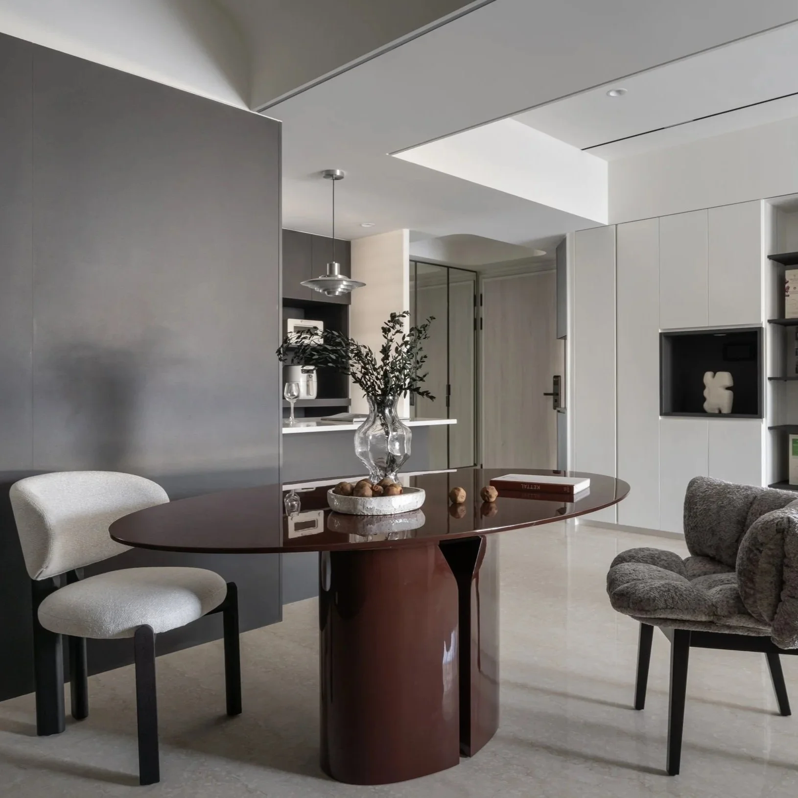 Modern living room and kitchen area with a round wooden table, white upholstered chair, gray sofa, decorative vase with greenery, books, and built-in white cabinetry.