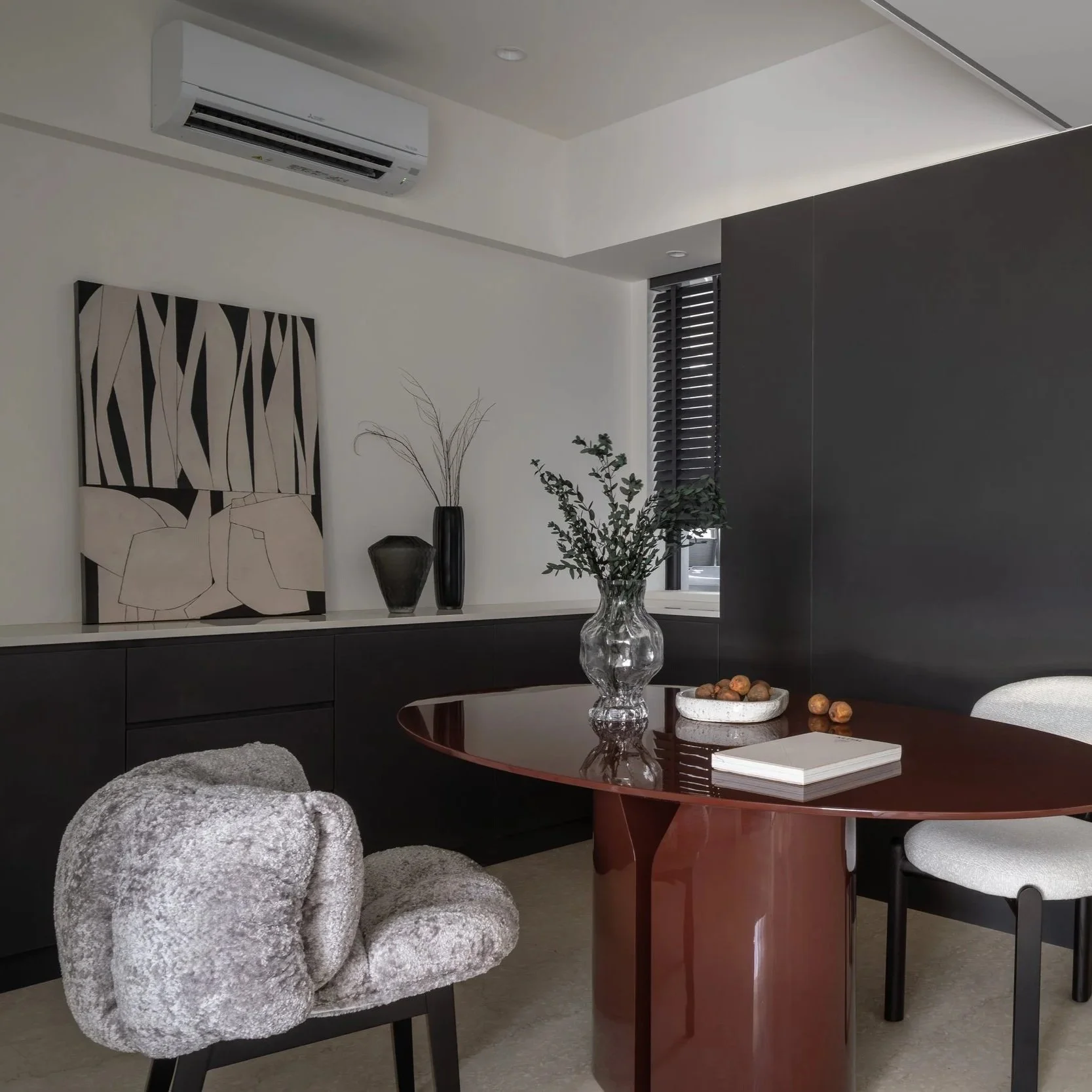 Interior of a modern dining room with a dark wood dining table, white upholstered chairs, a decorative black and white artwork on the wall, a vase with greenery, a round white bowl with nuts, and a window with black blinds. An air conditioning unit i