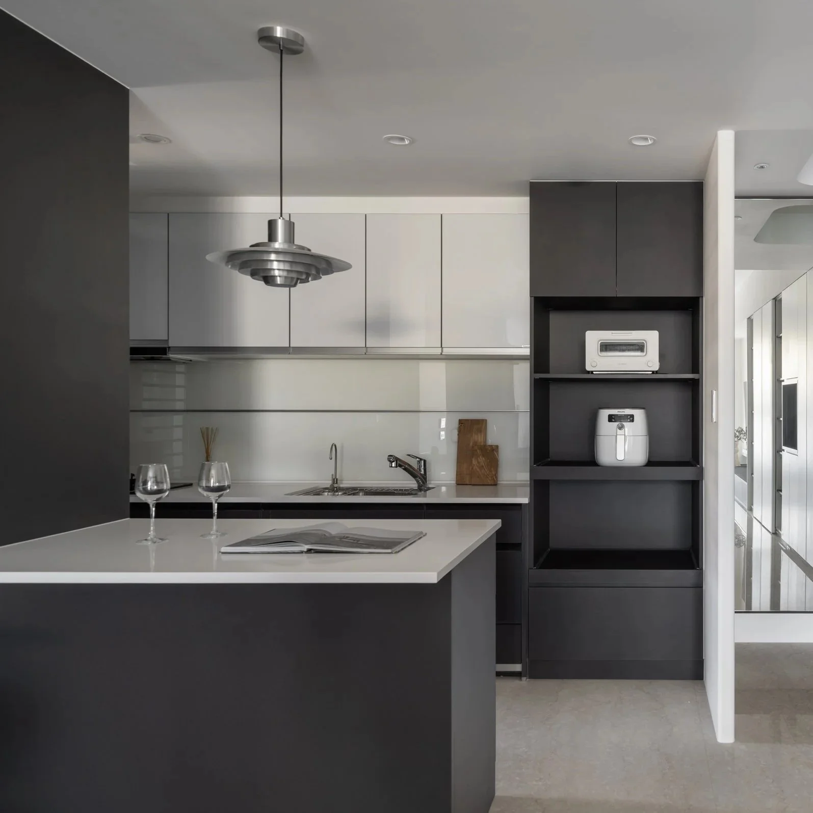 Modern kitchen with white countertops, gray and black cabinets, a silver hanging light fixture, and two wine glasses on a counter in the foreground. A bread toaster and rice cooker are visible on a black open shelving unit.