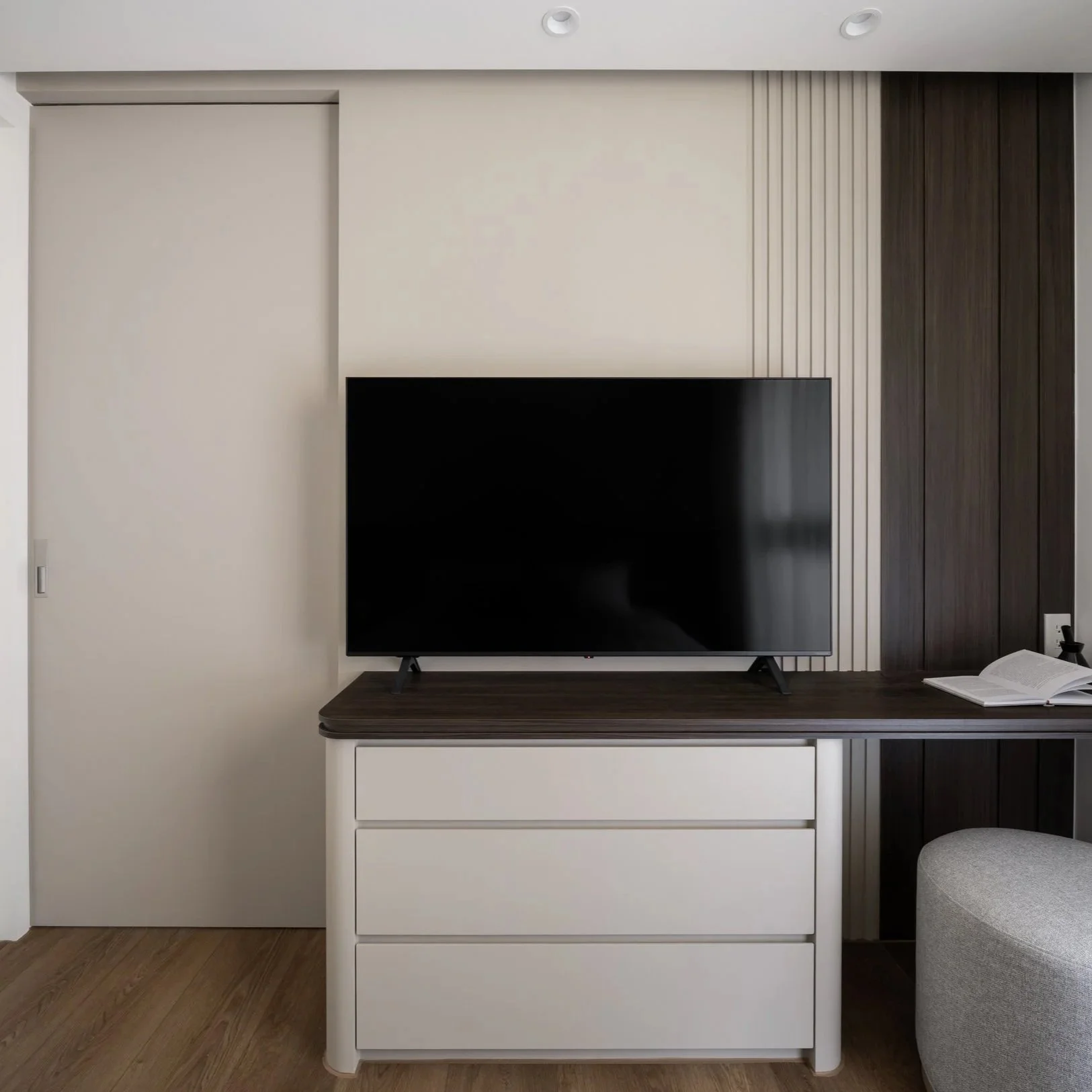 Living room with a black flat-screen TV on a dark wood table, a white dresser with four drawers, and a gray upholstered ottoman. The background wall has a beige section with vertical wood slats and dark wood panels on the right side.