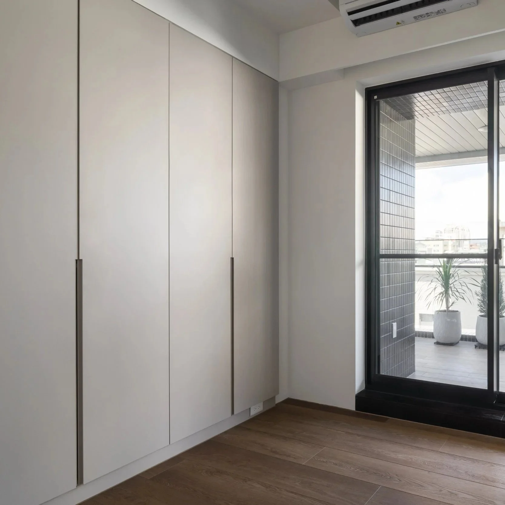 Interior view of a modern apartment or office with white built-in cabinets, a sliding glass door leading to a balcony or terrace, and a potted plant outside.