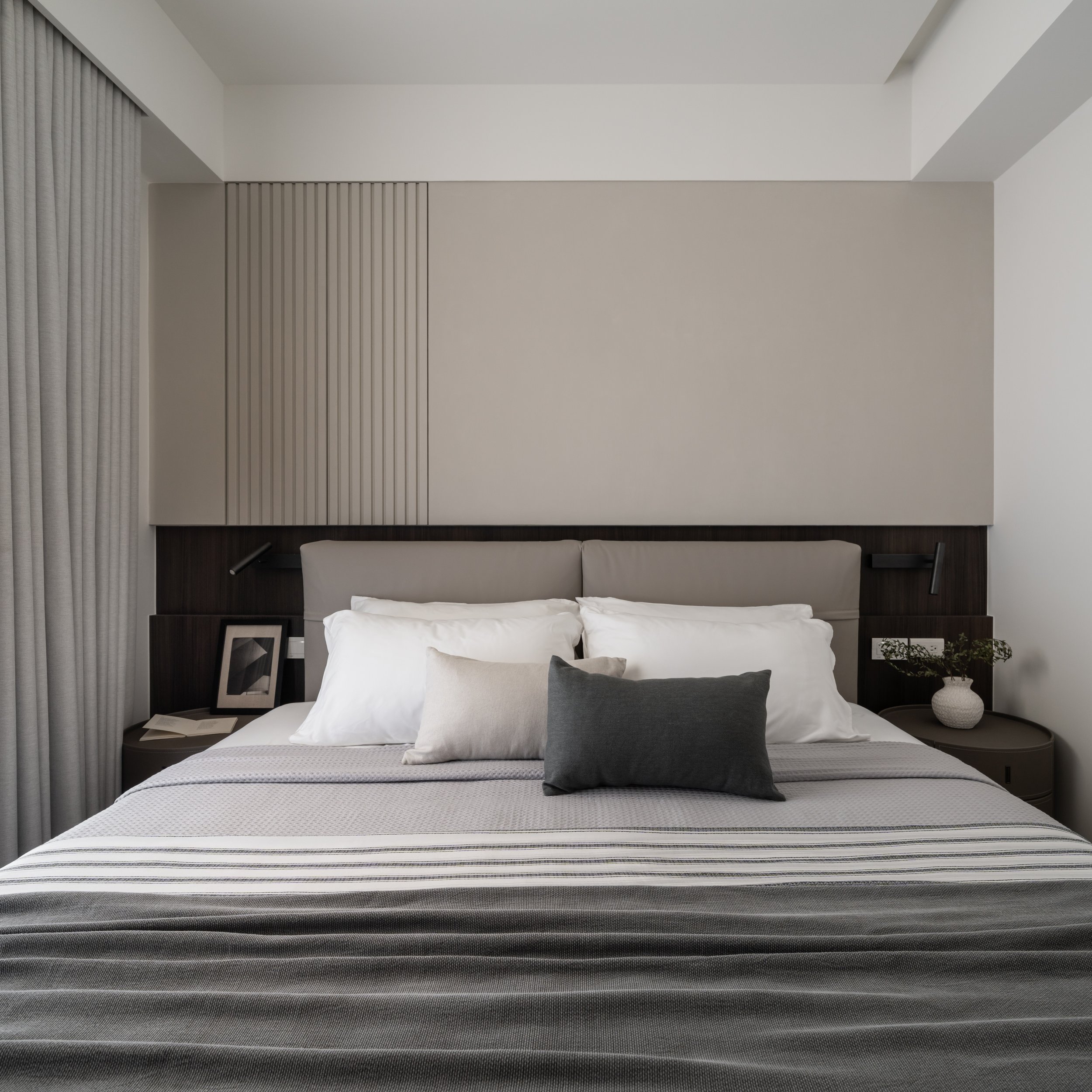 Modern bedroom with a large bed, multiple pillows, and a headboard featuring vertical wood slats. The room has a neutral color palette, with a framed photograph on the nightstand and a decorative vase with greenery. Curtains are drawn to the side ove