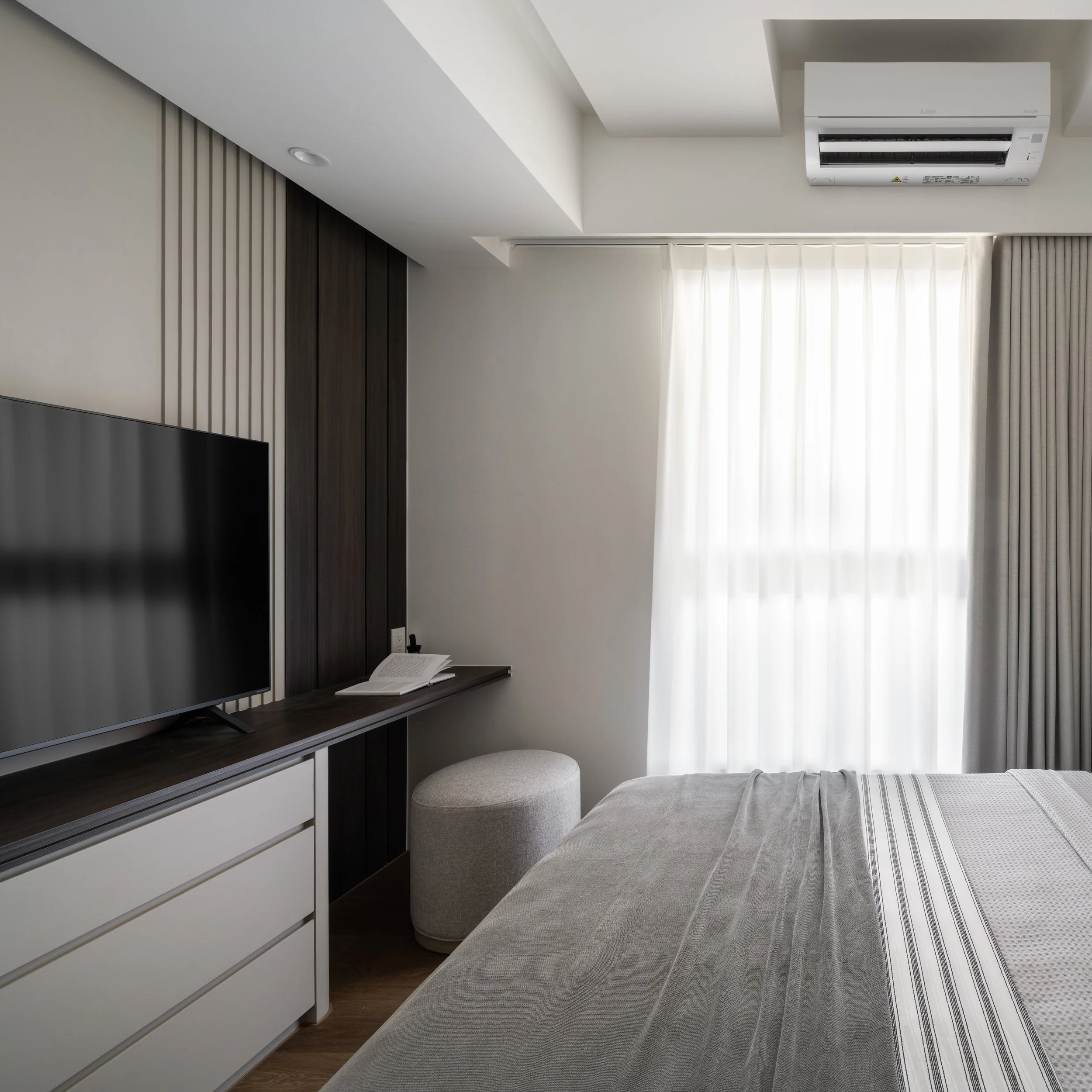 A modern living room corner showing a wall-mounted TV, a dark wood wall panel, a light-colored sofa, a small round ottoman, a window with white curtains, a book on a dark wood shelf, and a ceiling-mounted air conditioner.