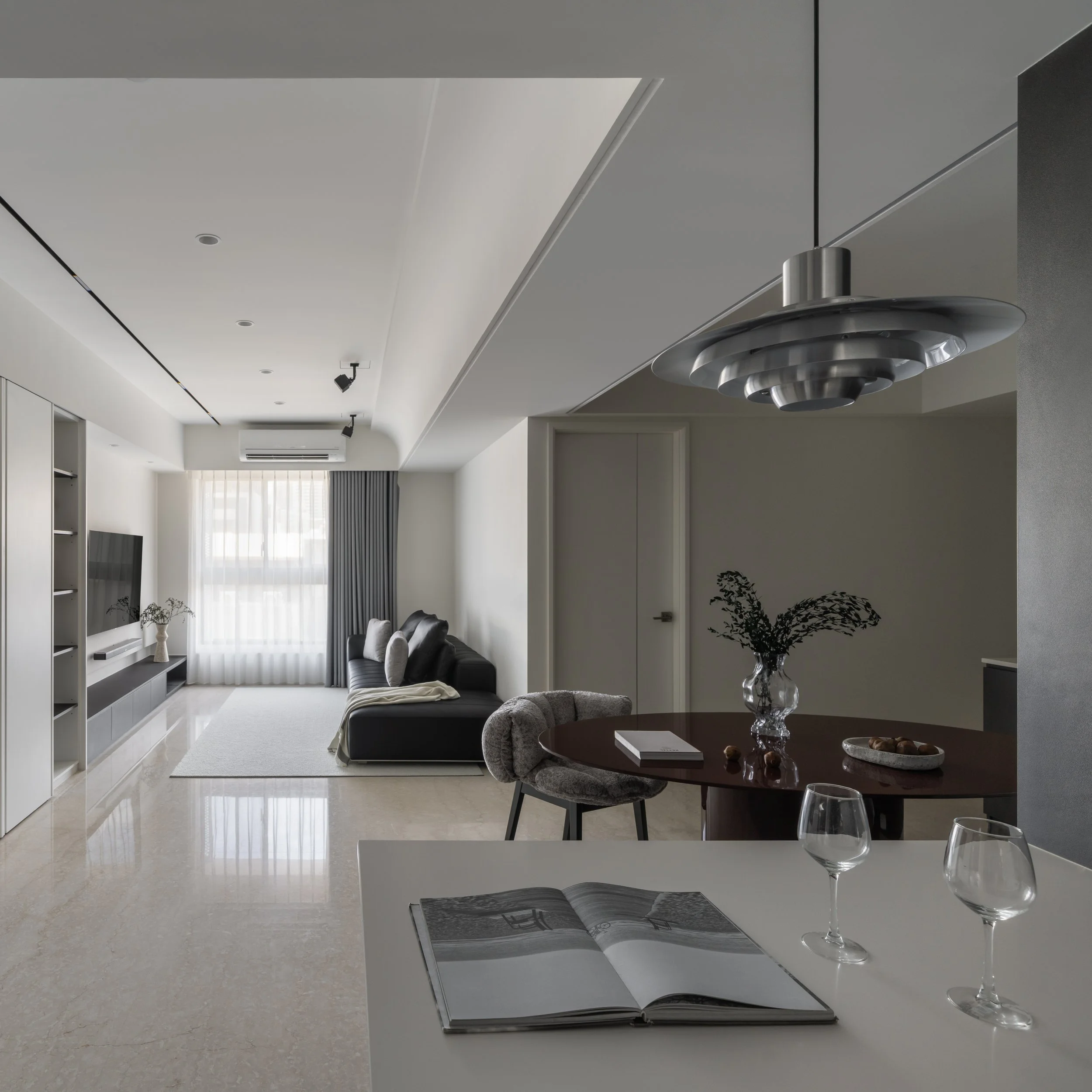 Modern living room with white walls, gray curtains, a black sofa with pillows, a white coffee table, and a large window with sheer curtains letting in natural light.