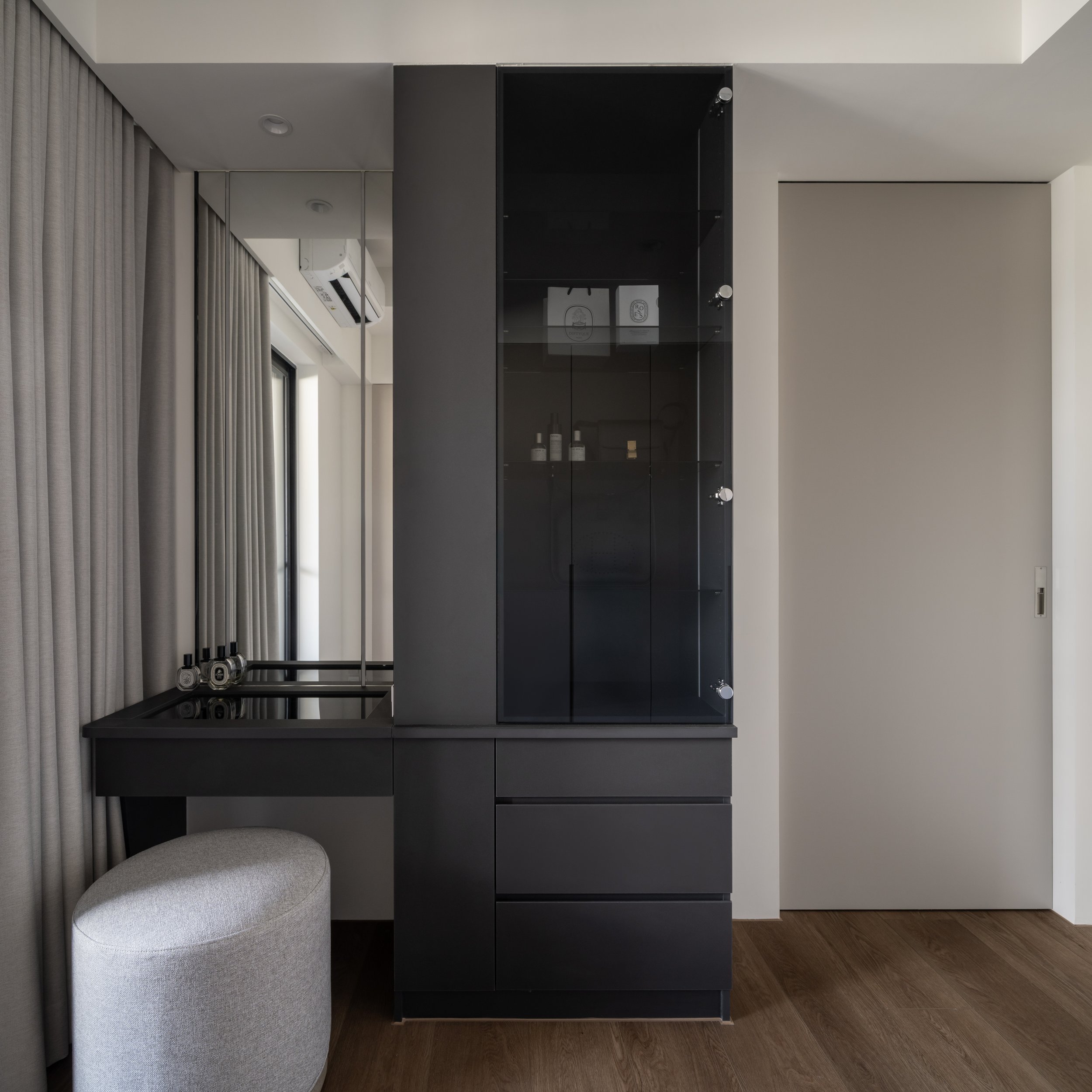 Modern black display cabinet with glass doors, two shelves, and some products inside, next to a gray armchair and beige curtains in a minimalist room.